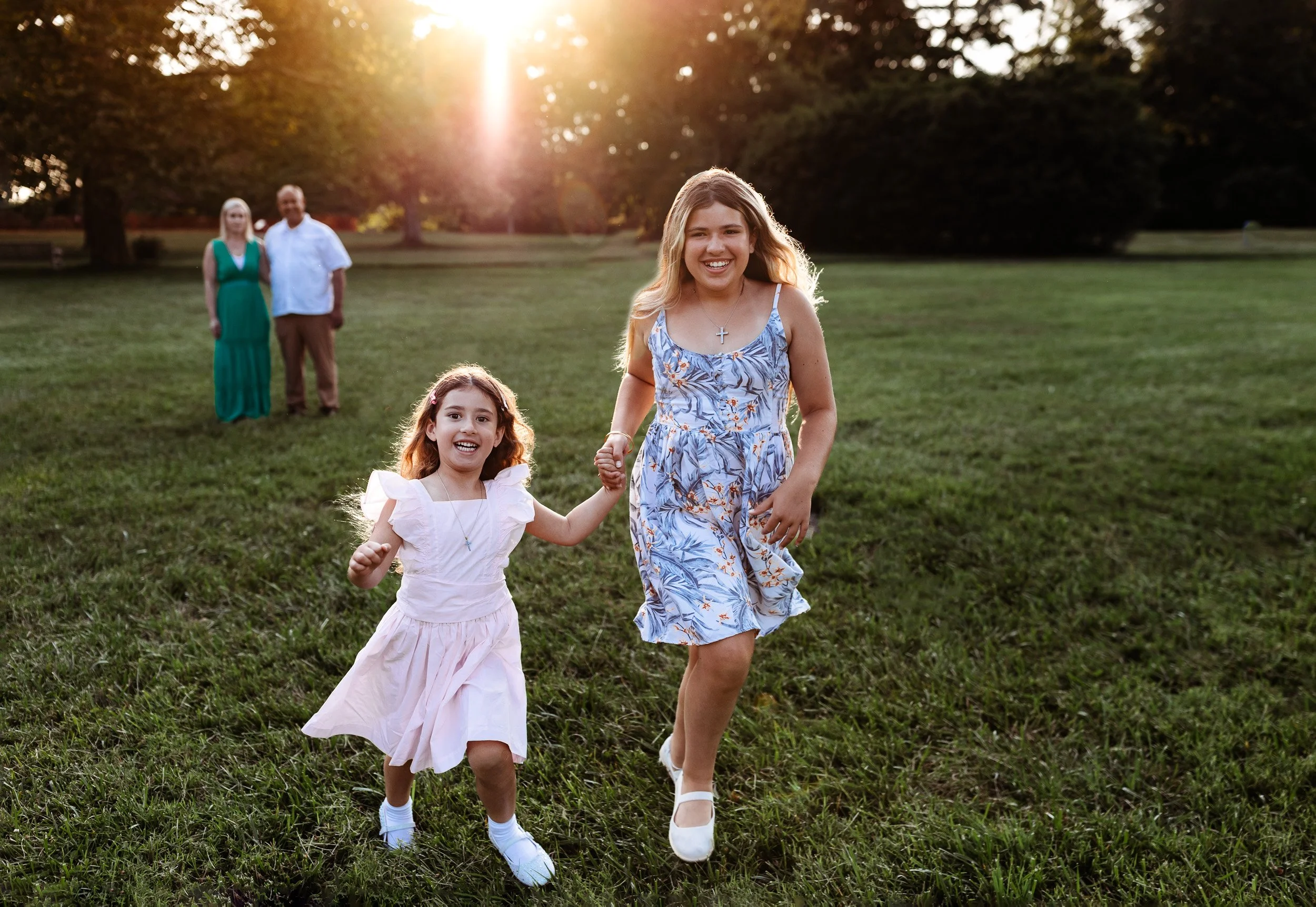 sisters running a field at sunset and they are laughing while parents are in the background laughing