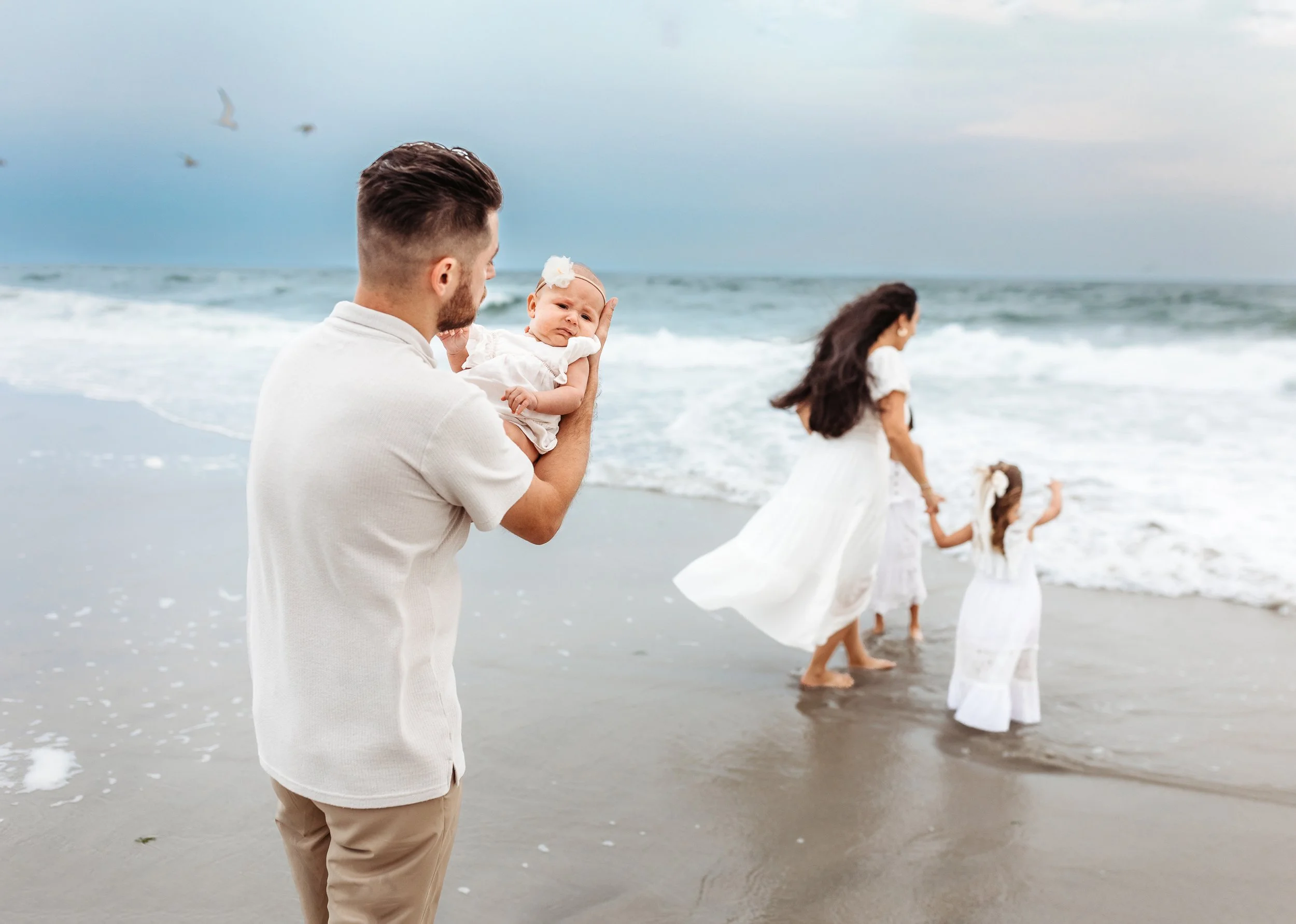 dad holding newborn at the ocean shore while mom is in background playing with older sisters