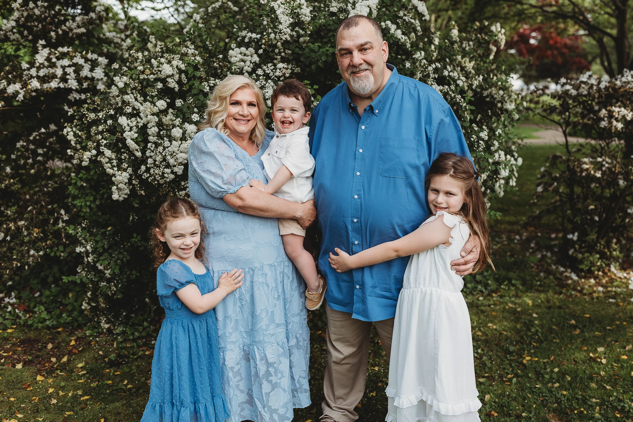 grandparents with their grandchildren hugging them.  dressed in blue and white in front to a pretty floral bush 