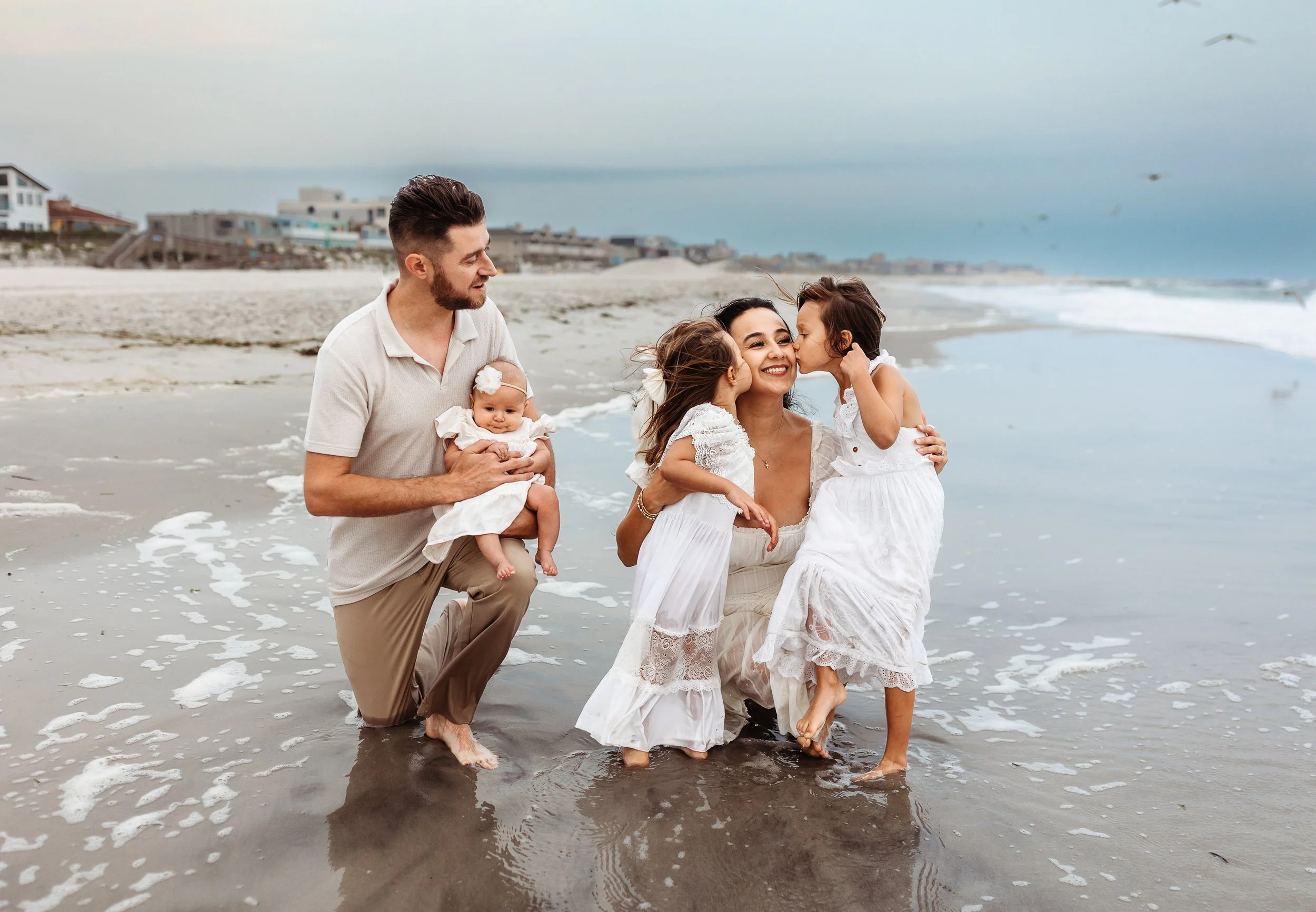 sisters kissing mom in the waves while dad kneels holding baby and watches