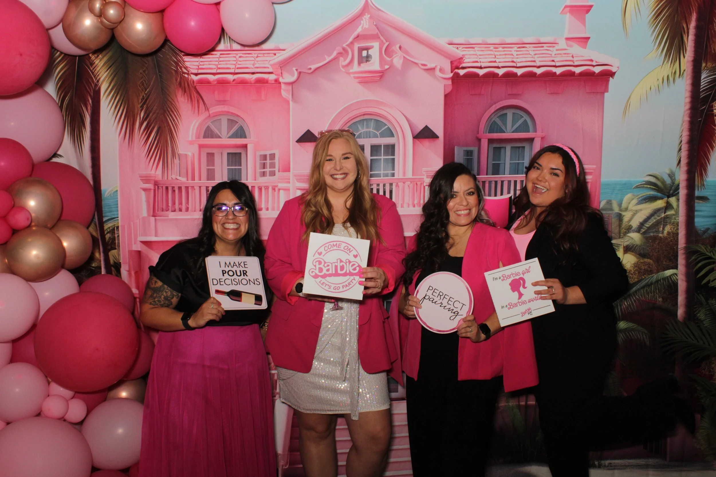 Four women at a pink-themed party with a Barbie house backdrop, holding signs with playful messages.