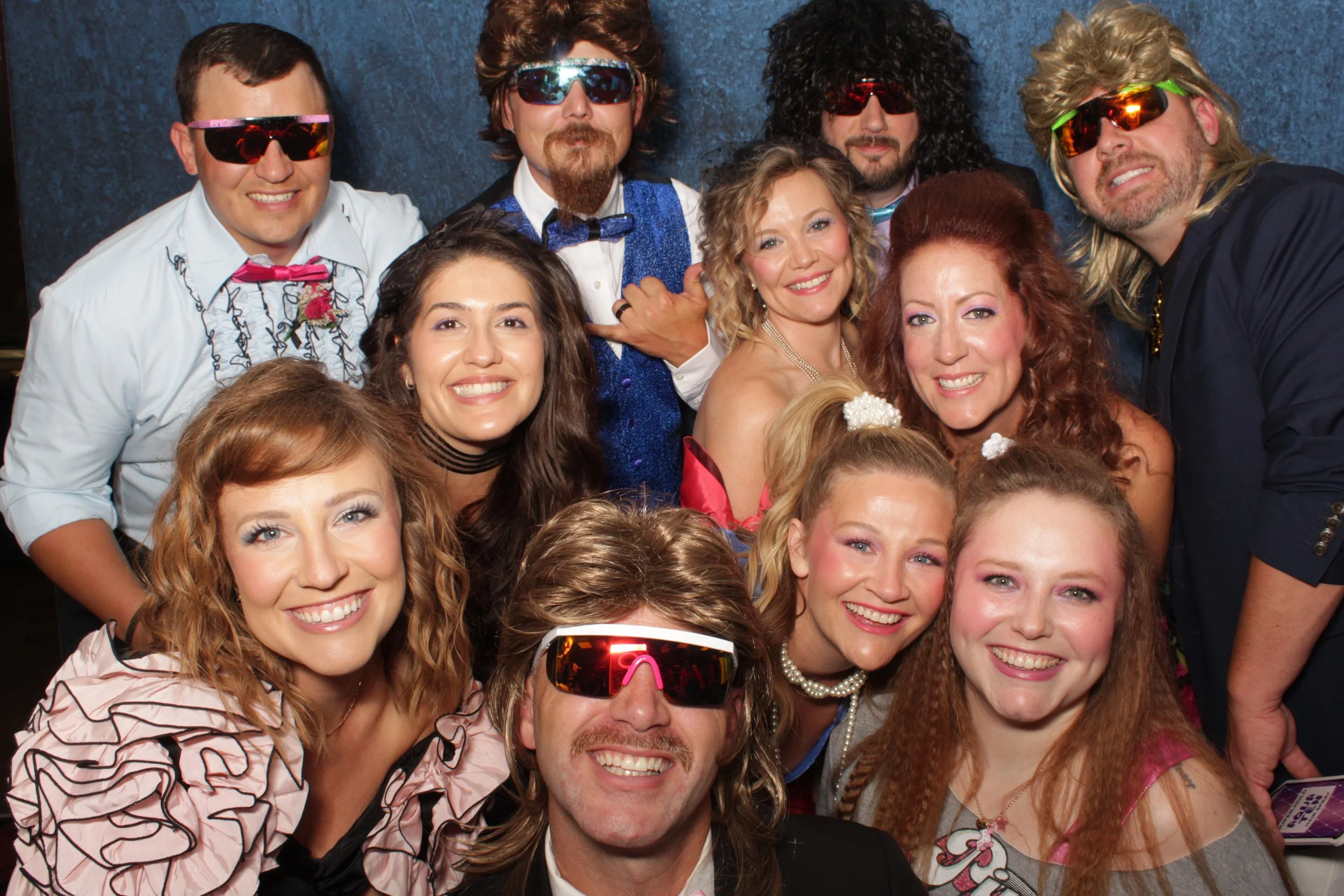 Group of people dressed in 80s costumes and sunglasses, smiling and taking a selfie.