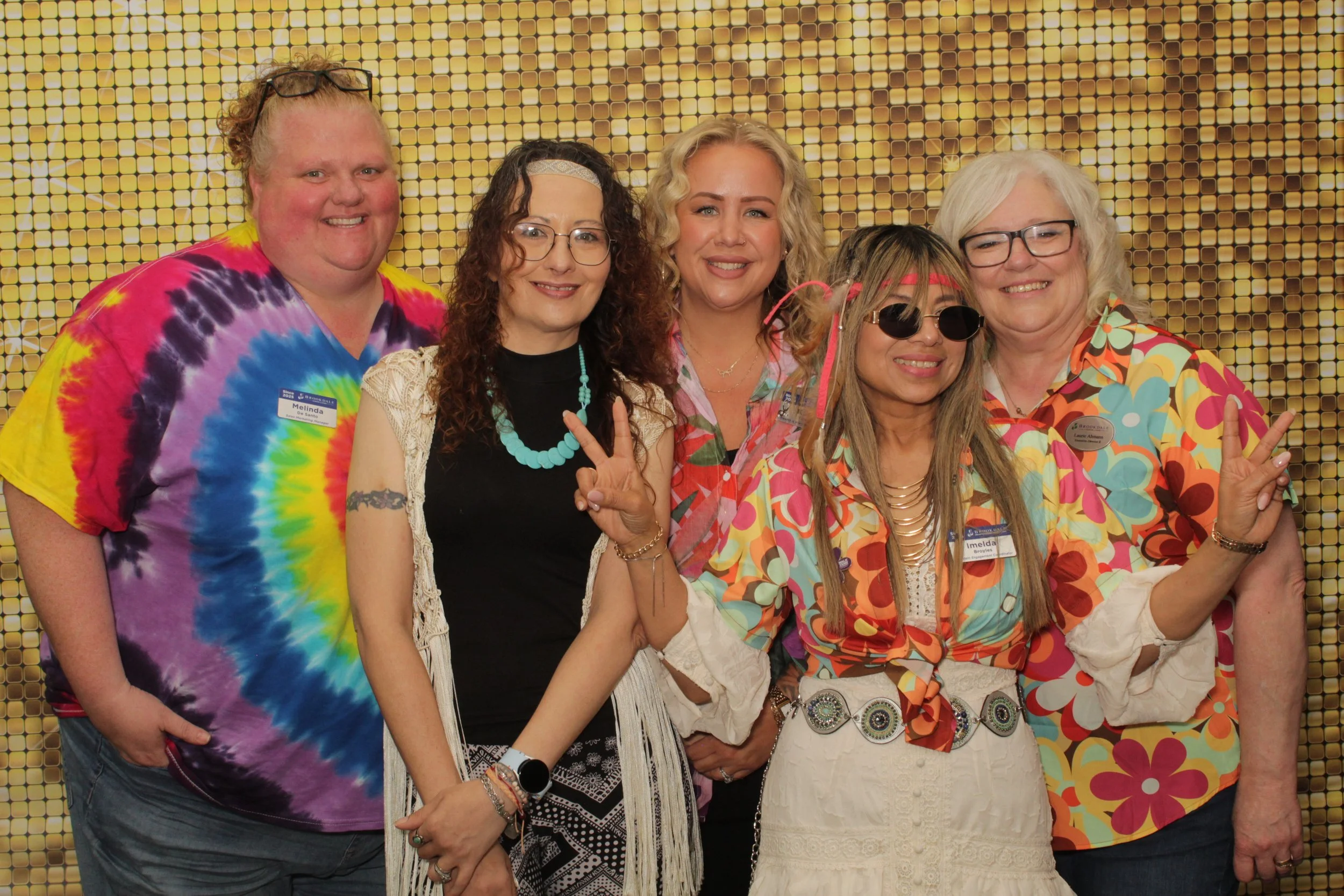 Six women standing together, smiling, in front of a gold backdrop. They are wearing colorful, retro, 1960s and 1970s style clothing and accessories, making peace signs and peace symbols with their hands.