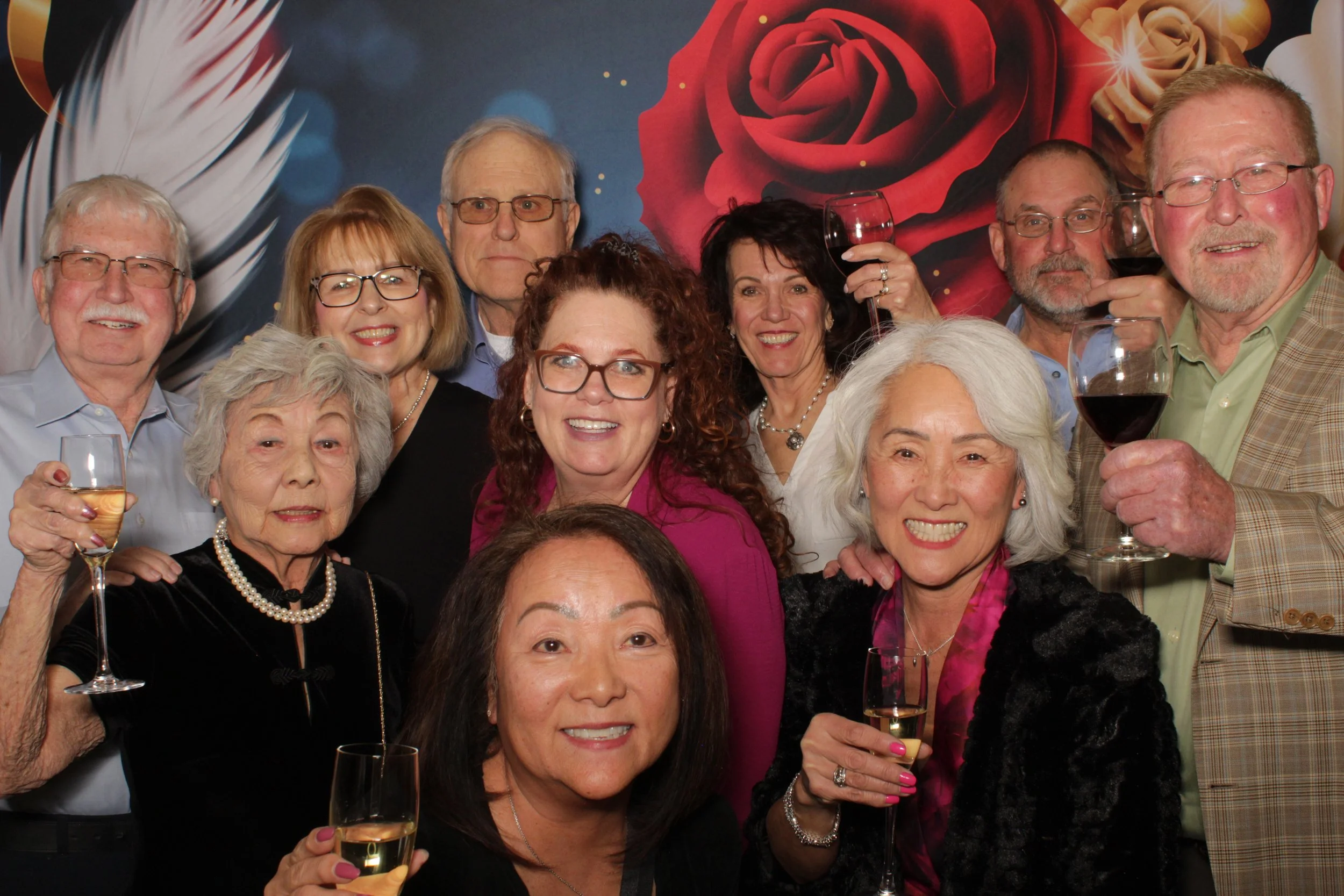 Group of eleven older adults celebrating with wine glasses, smiling, in front of a colorful floral backdrop.