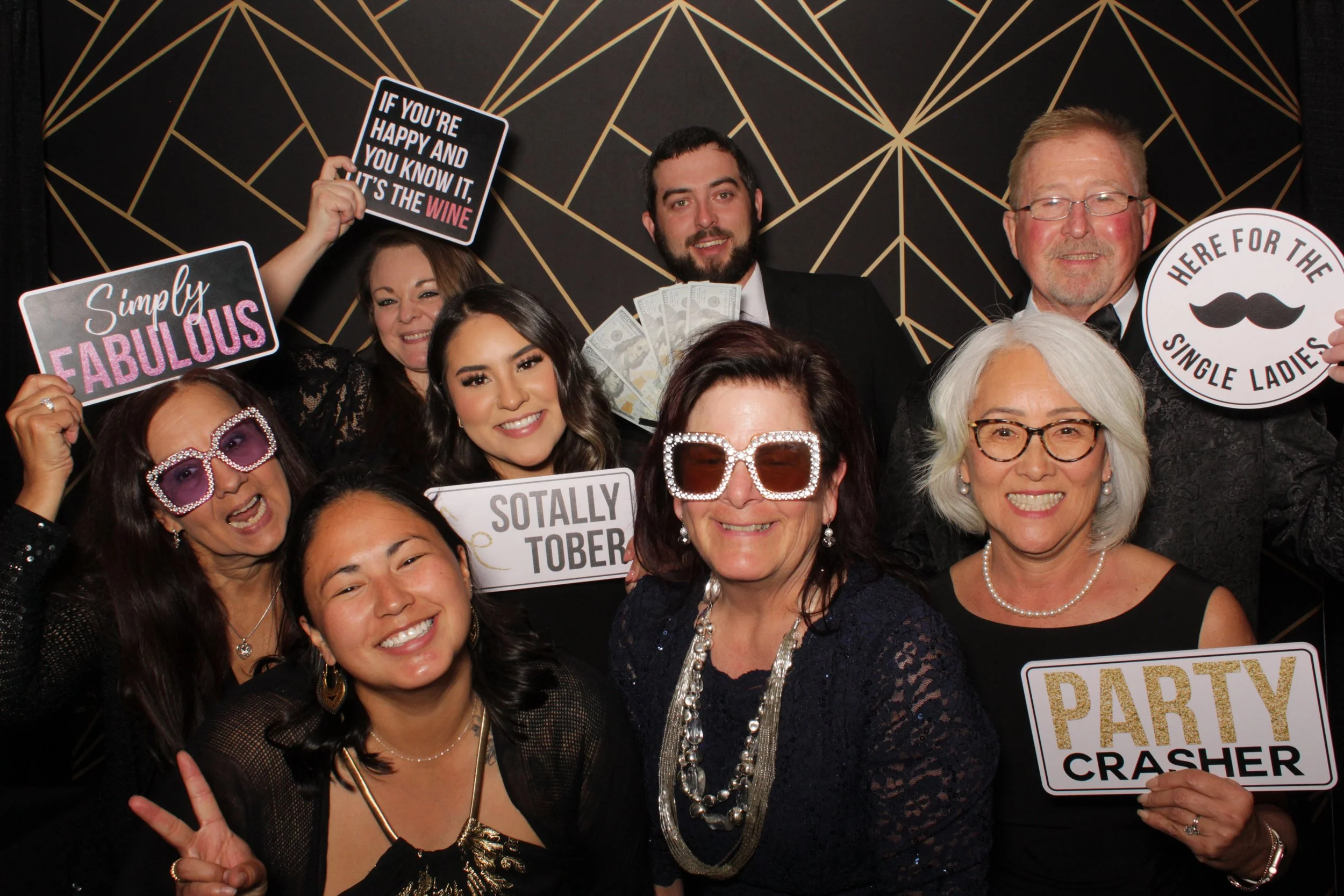 Group of nine people at a party, holding signs and wearing fun accessories, in front of a black backdrop with gold line patterns.