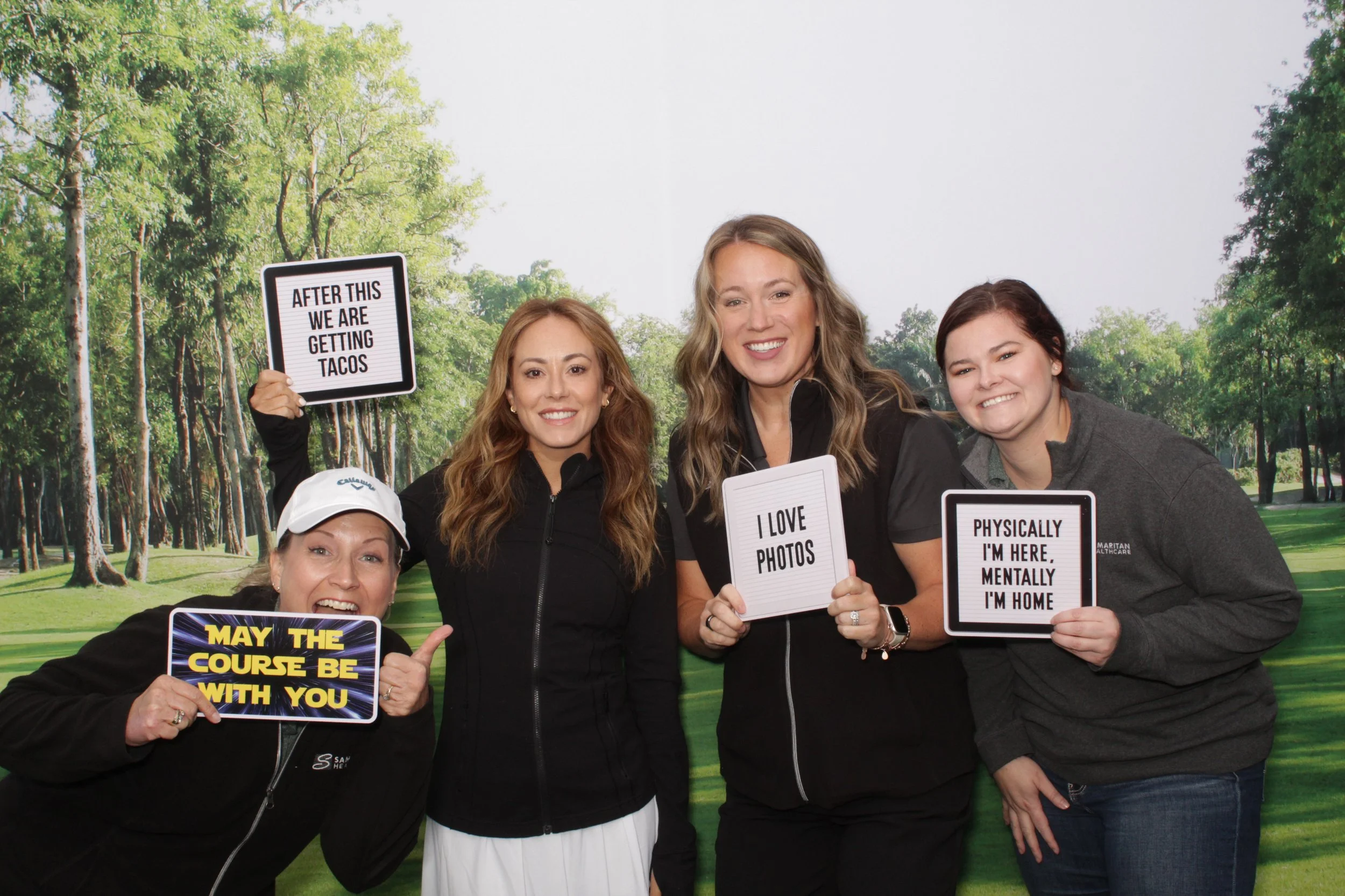 Four smiling women holding signs at a golf course, with trees in the background. The signs read: 'AFTER THIS WE ARE GETTING TACOS', 'MAY THE COURSE BE WITH YOU', 'I LOVE PHOTOS', and 'PHYSICALLY I'M HERE, MENTALLY I'M HOME'.