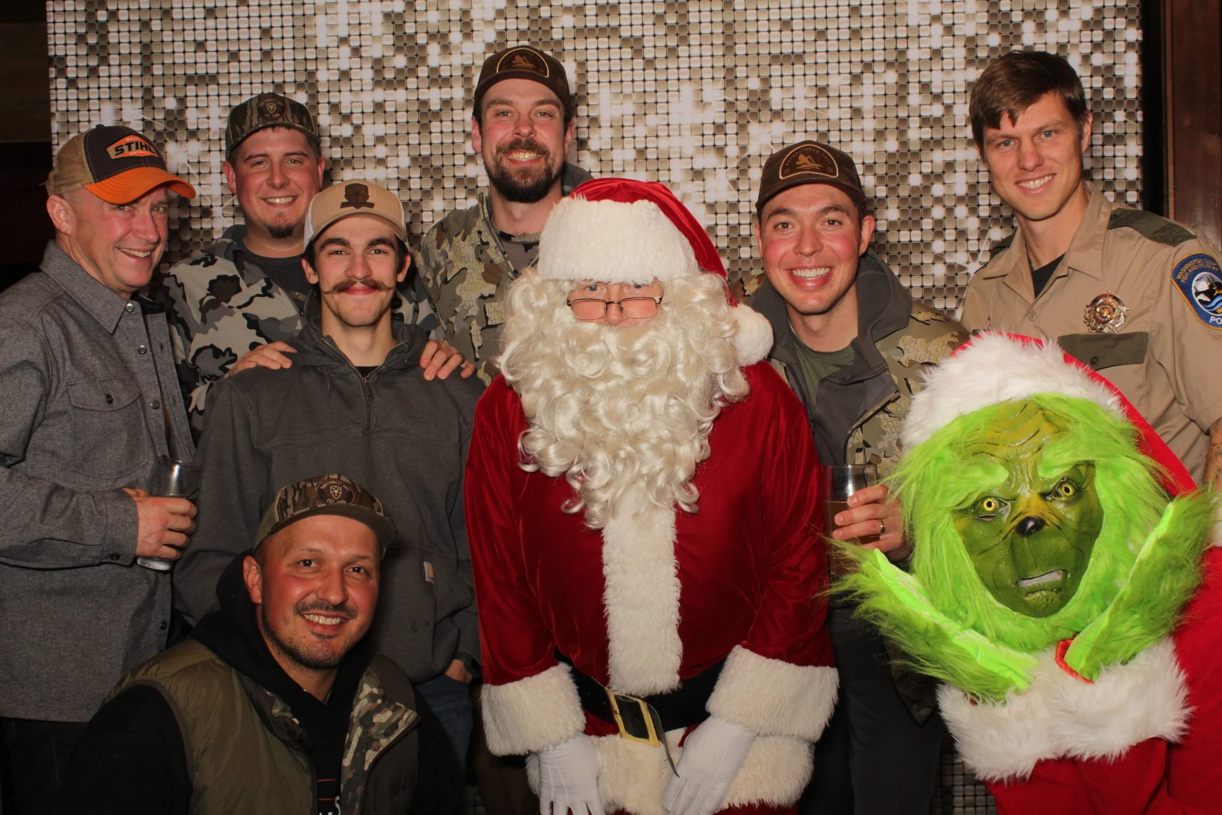 A group of nine people dressed in casual and camouflage clothing, standing with Santa Claus and the Grinch in front of a shiny backdrop. The group is smiling and holding drinks, appearing to celebrate a holiday event.