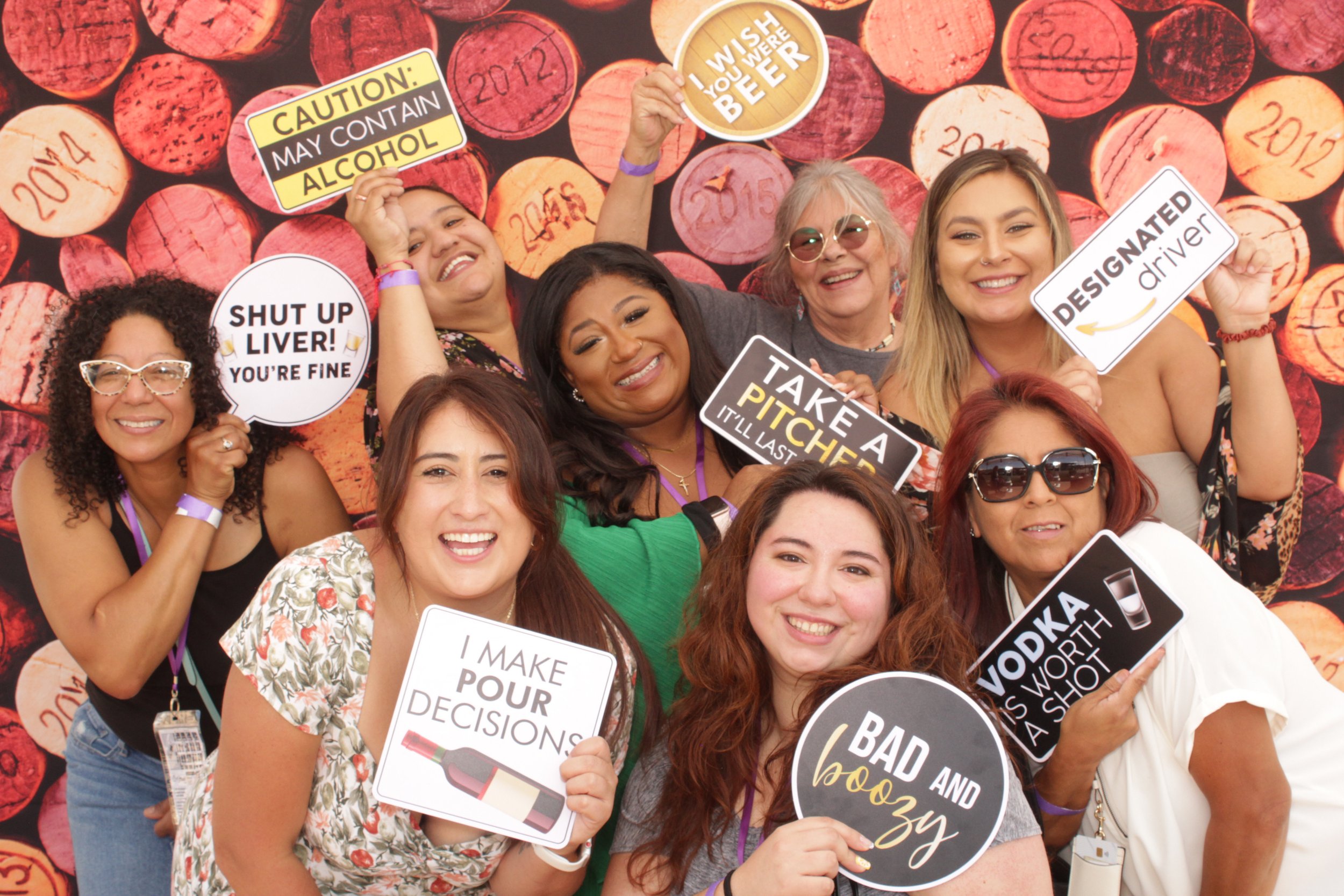 Group of women smiling and holding humorous signs with wine and alcohol references in front of a background of wine corks.