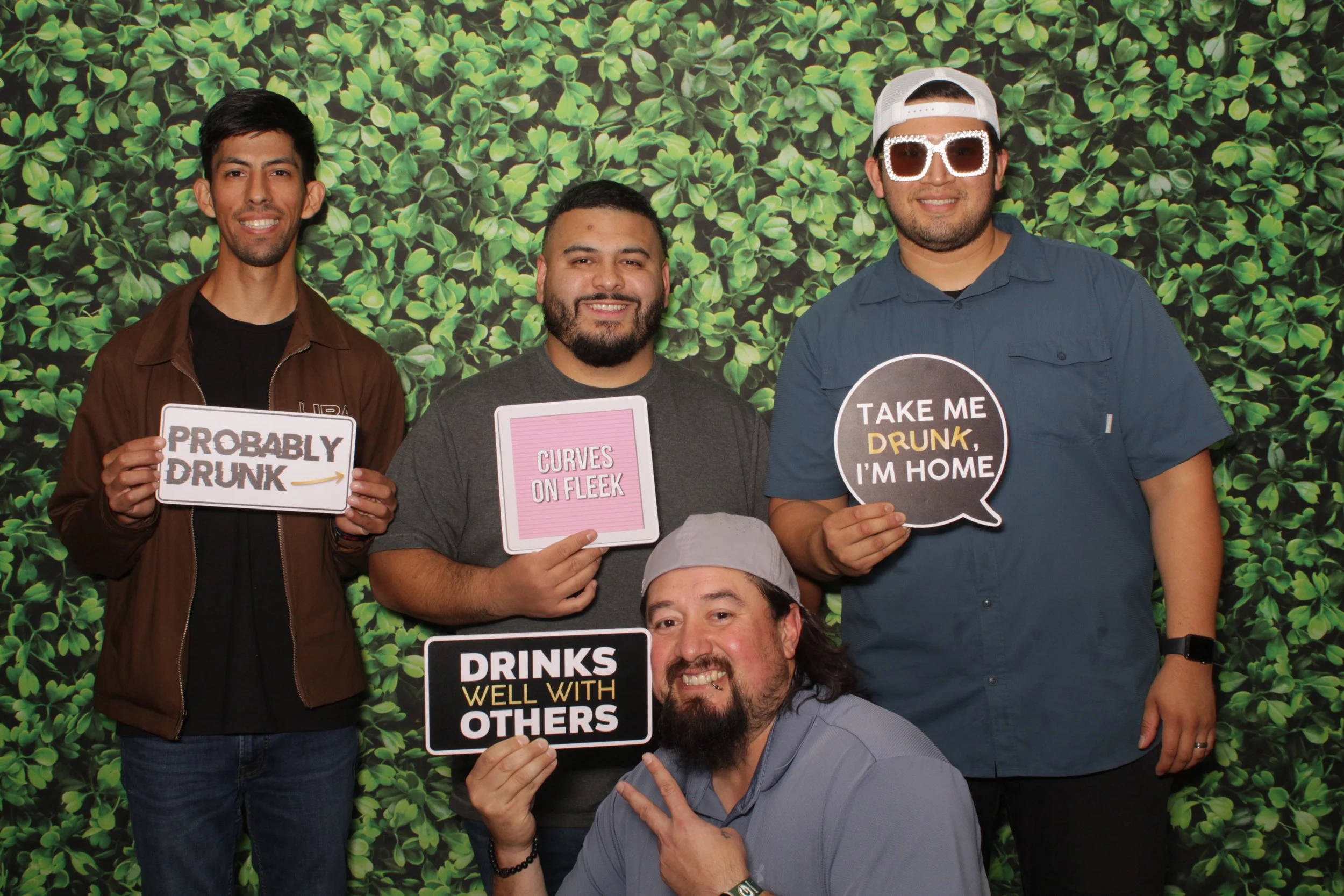 Four men posing in front of a leafy green backdrop, holding signs with humorous messages about drinking and relationships, with one man making a peace sign.