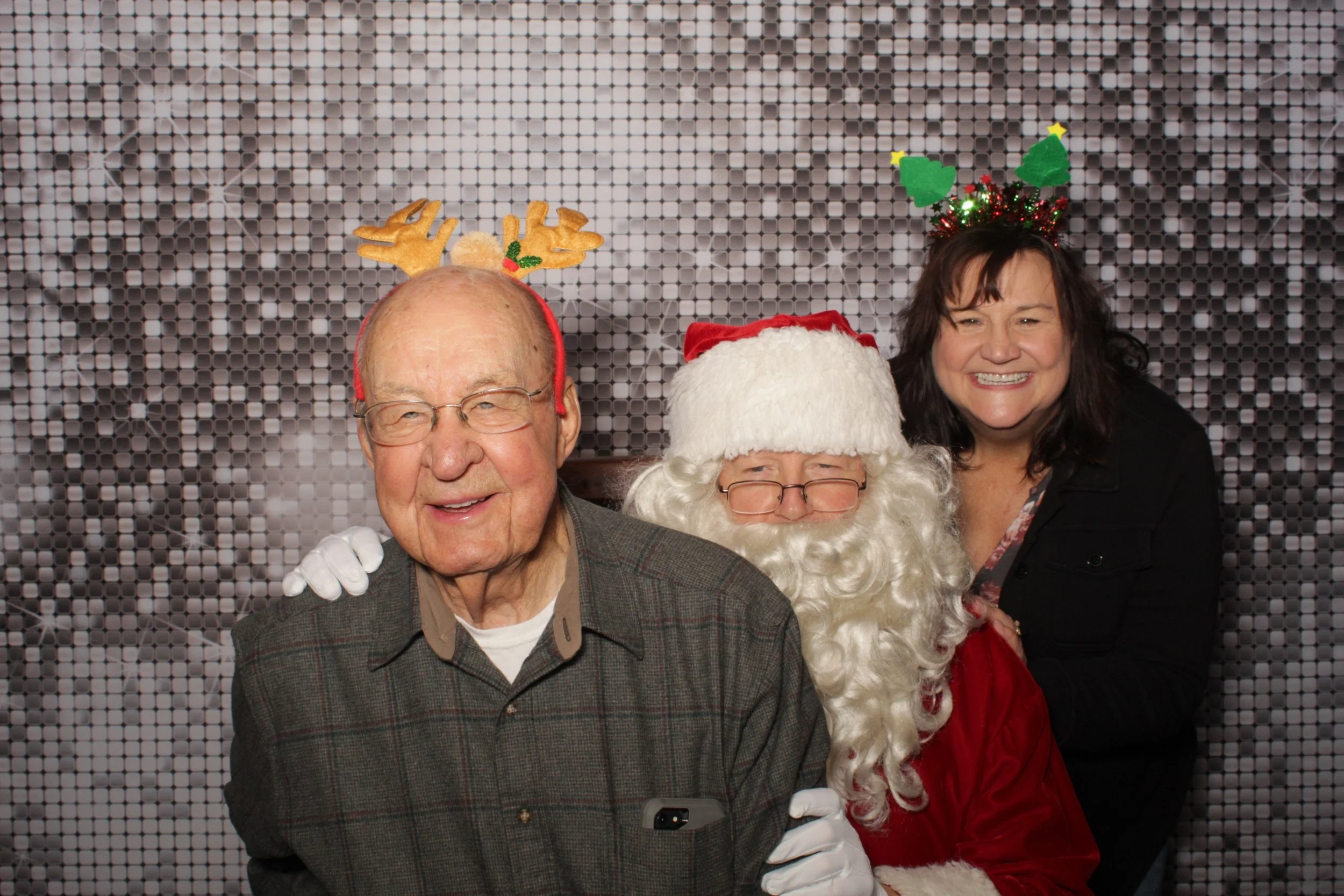 Group of three smiling people dressed in Christmas costumes. One man with reindeer antlers, a person dressed as Santa Claus, and a woman with a Christmas headband with wreaths.