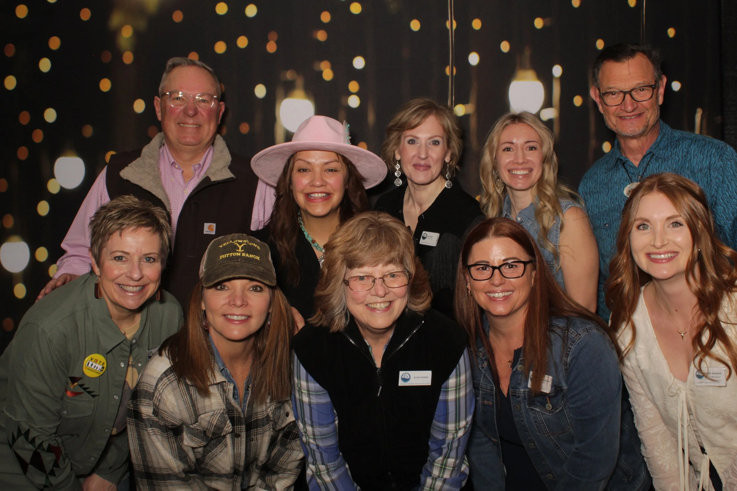 A group of eleven people, most with name tags, smiling at a photo event with a dark background of blurred lights.