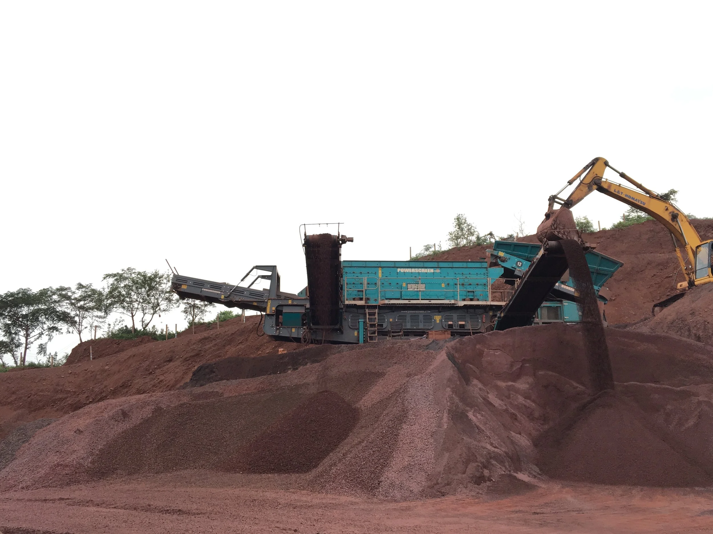 Excavator loading soil into a Powerscreen mobile crusher and conveyor at a construction site.