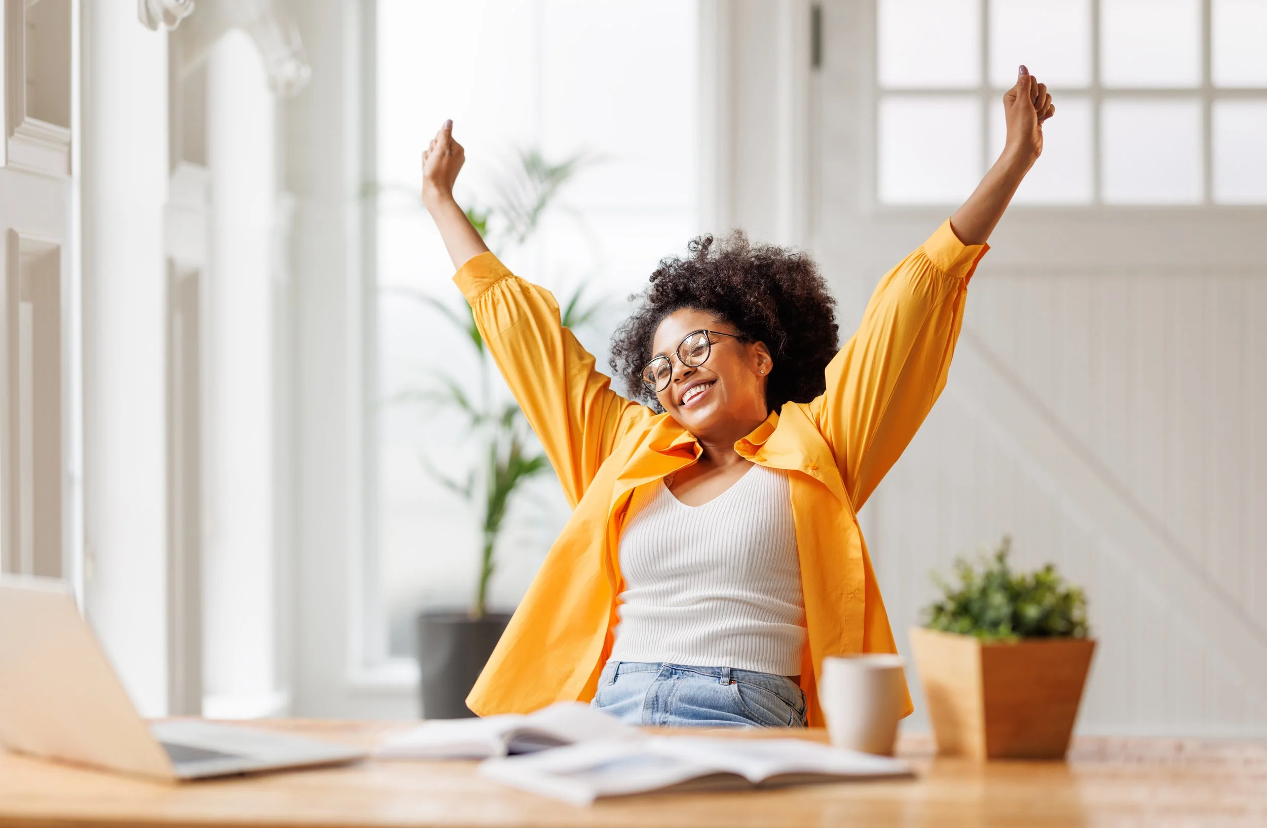 A happy woman with curly hair and glasses stretching with arms raised at her desk in a bright, modern home office.