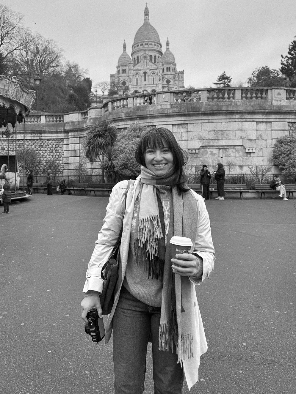 A woman, Bex Falk, smiling and holding a coffee cup in front of the Sacré-Cœur Basilica in Paris, France, with a carousel and park area in the background, in black and white.