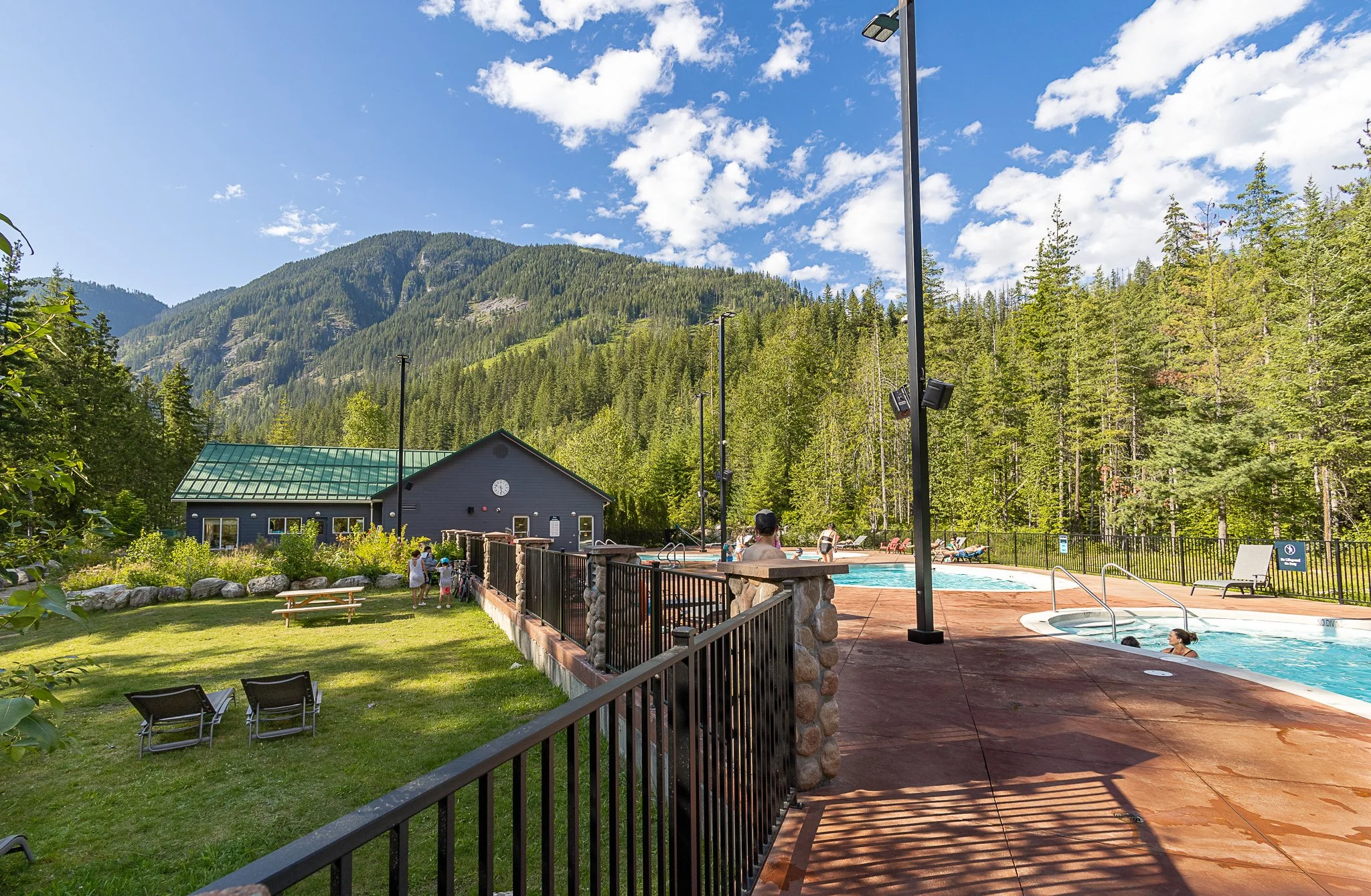 Guests relaxing in Crazy Creek's outdoor hot pools on a sunny summer day, with the lawn, pool building and a forested mountain behind