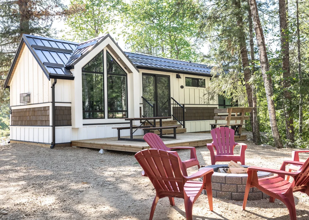 A white Crazy Creek cabin with large windows and a deck, with red chairs around a fire pit in the forest