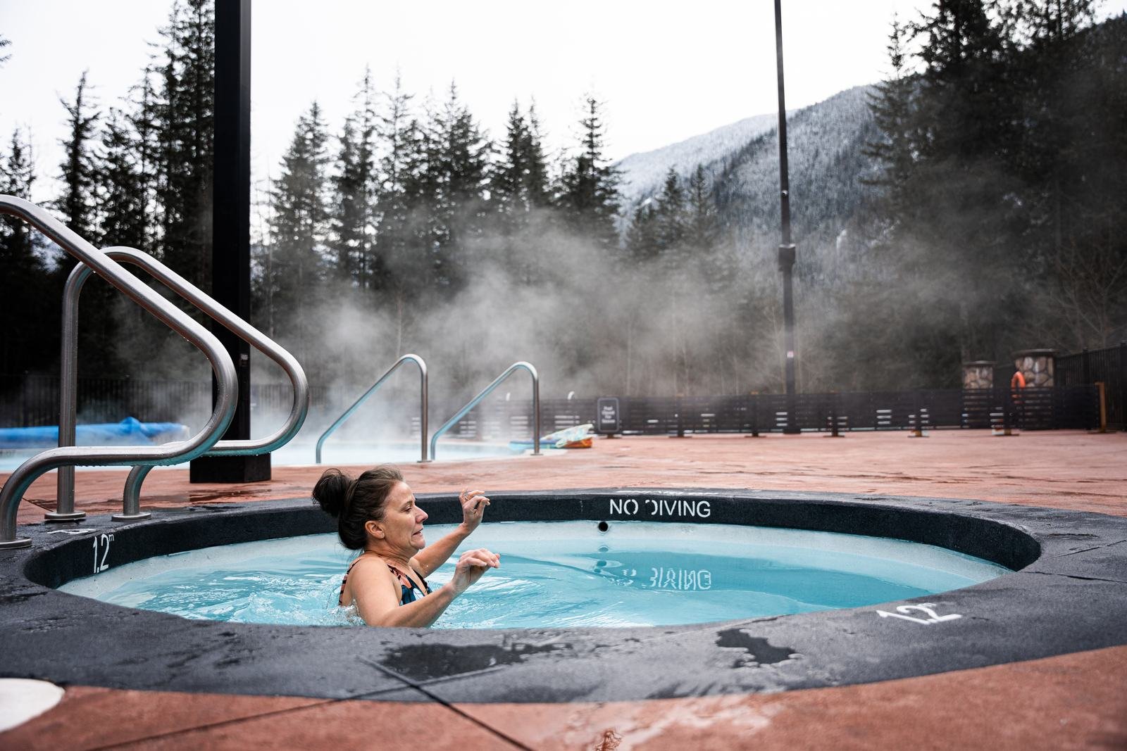 A woman easing into the cold plunge on a winter day, with steam drifting off the hot pools behind