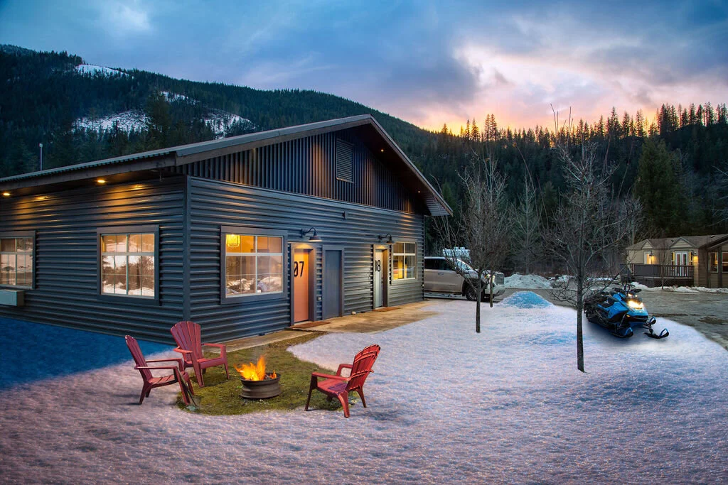 A Crazy Creek cabin in the snow at dusk with snowmobiles parked beside it and red chairs around the fire pit