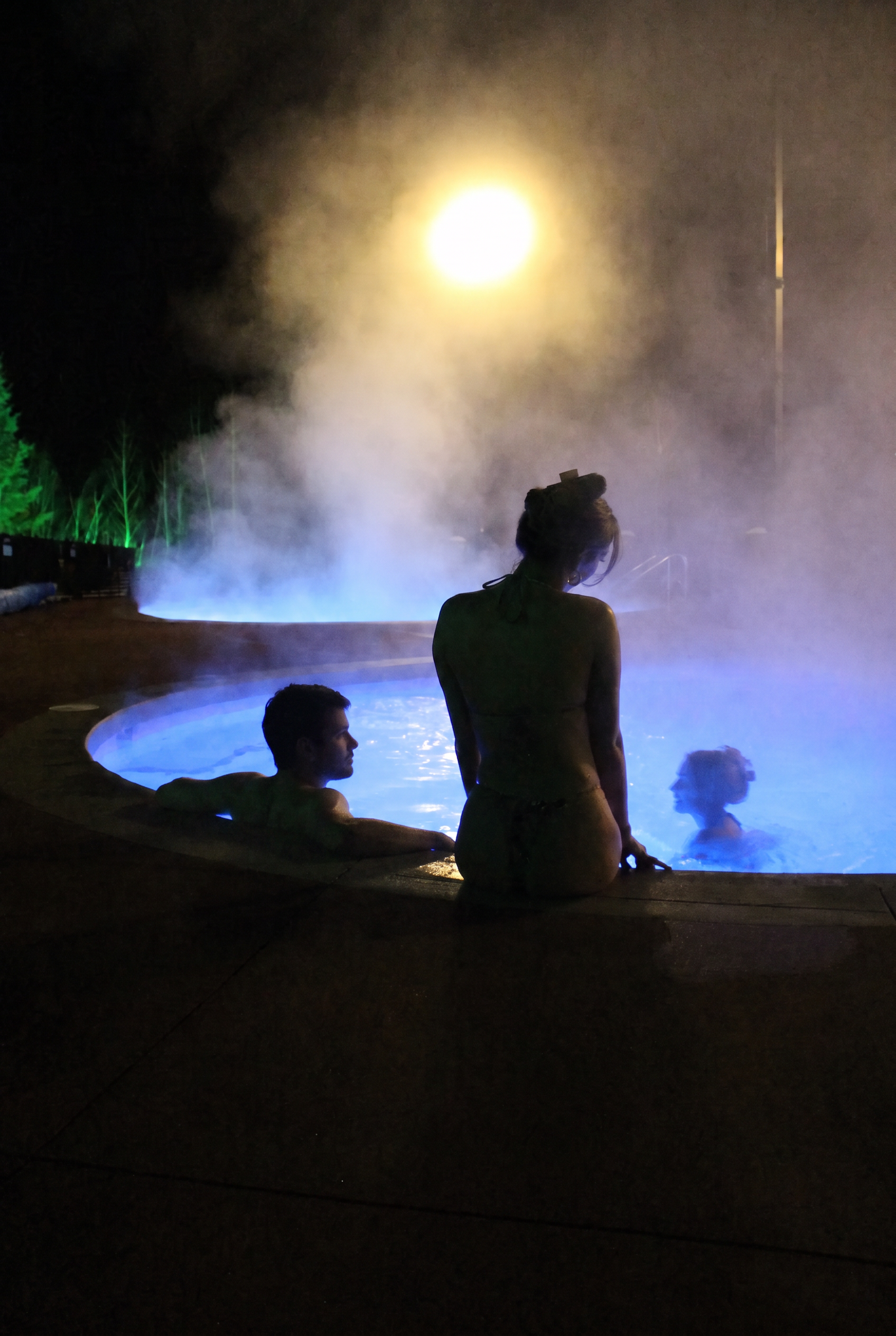 Nighttime scene at a hot spring with steam rising from the water, illuminated by colorful lights. Several people are relaxing in the hot spring, including a woman standing at the edge and others sitting in the water.