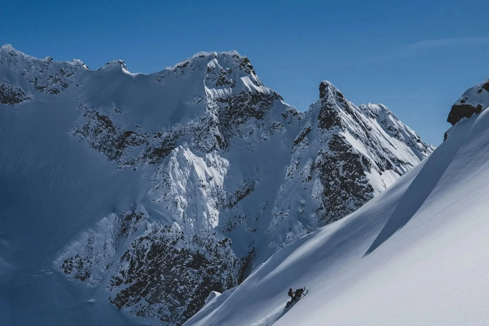 Snowmobiler riding a steep snowy slope below rugged mountain peaks