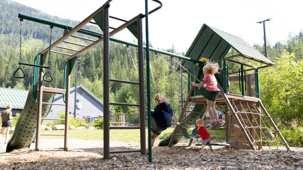 Kids playing on the Crazy Creek playground, with slides and climbing structures in front of a forested hill