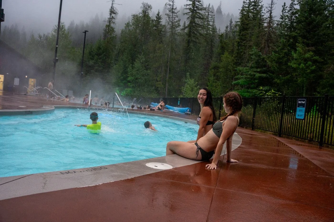 Two friends laughing on the rain-soaked pool deck beside a steaming pool, with misty forest behind