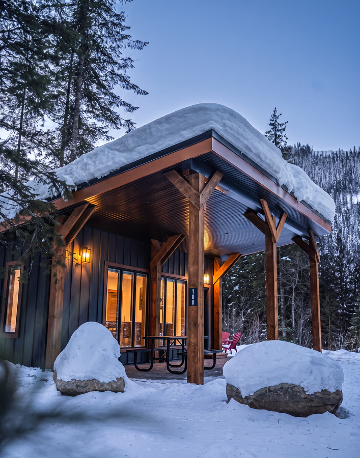 A cozy cabin in a snowy landscape, with warm lights glowing through the glass doors and windows, surrounded by snow-covered rocks and trees.