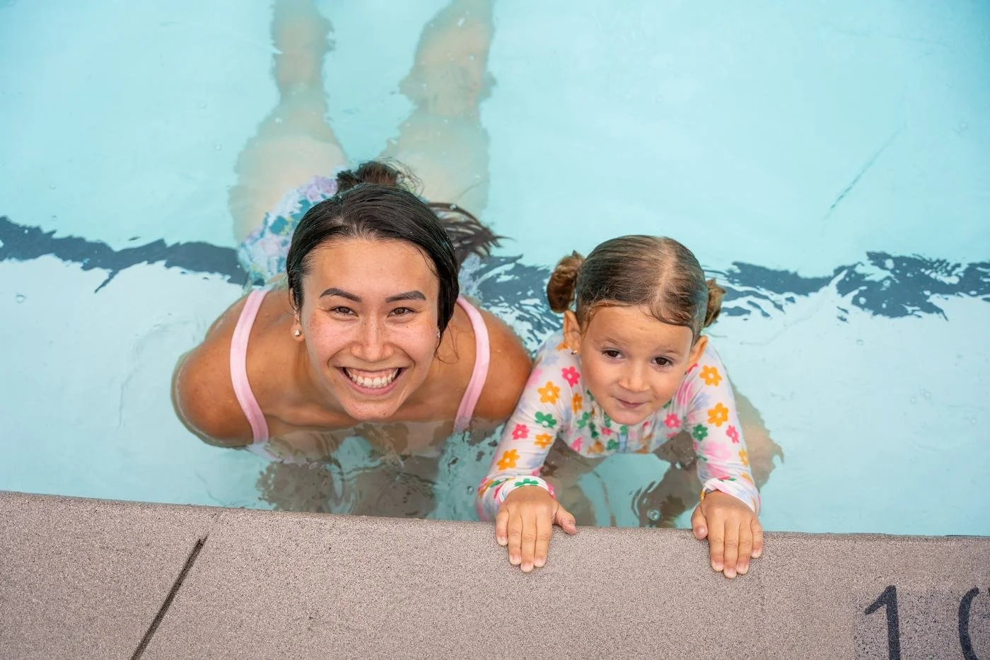 A mother and young daughter smiling up at the camera from the edge of a pool