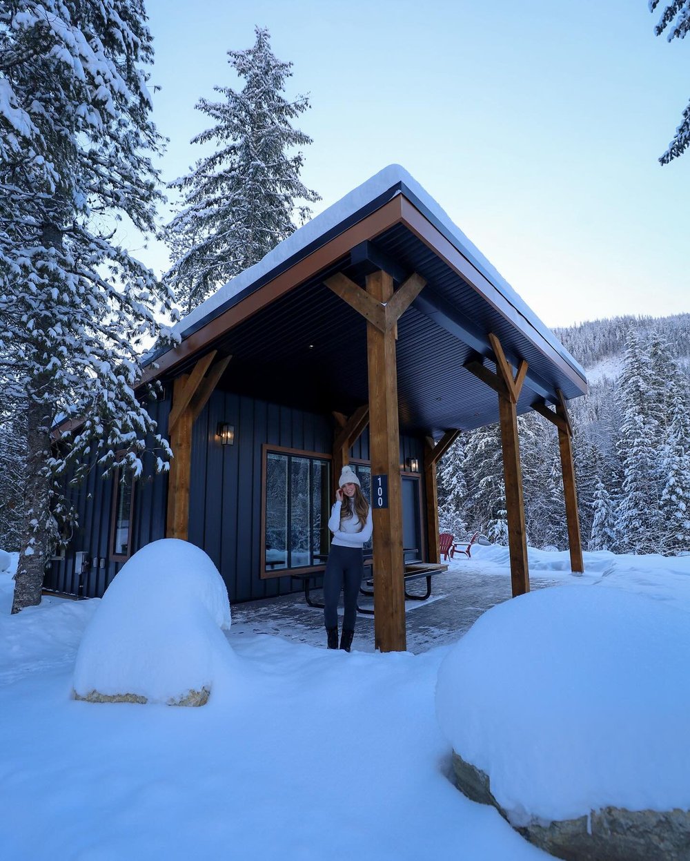 A guest in a toque on the covered porch of a dark forest cabin in deep winter snow
