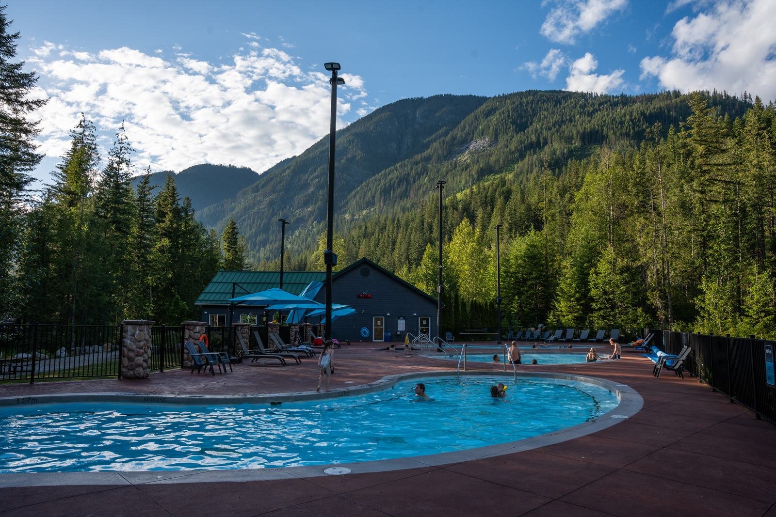 Guests soaking in a sunny outdoor pool at Crazy Creek, with forested mountains and blue sky behind