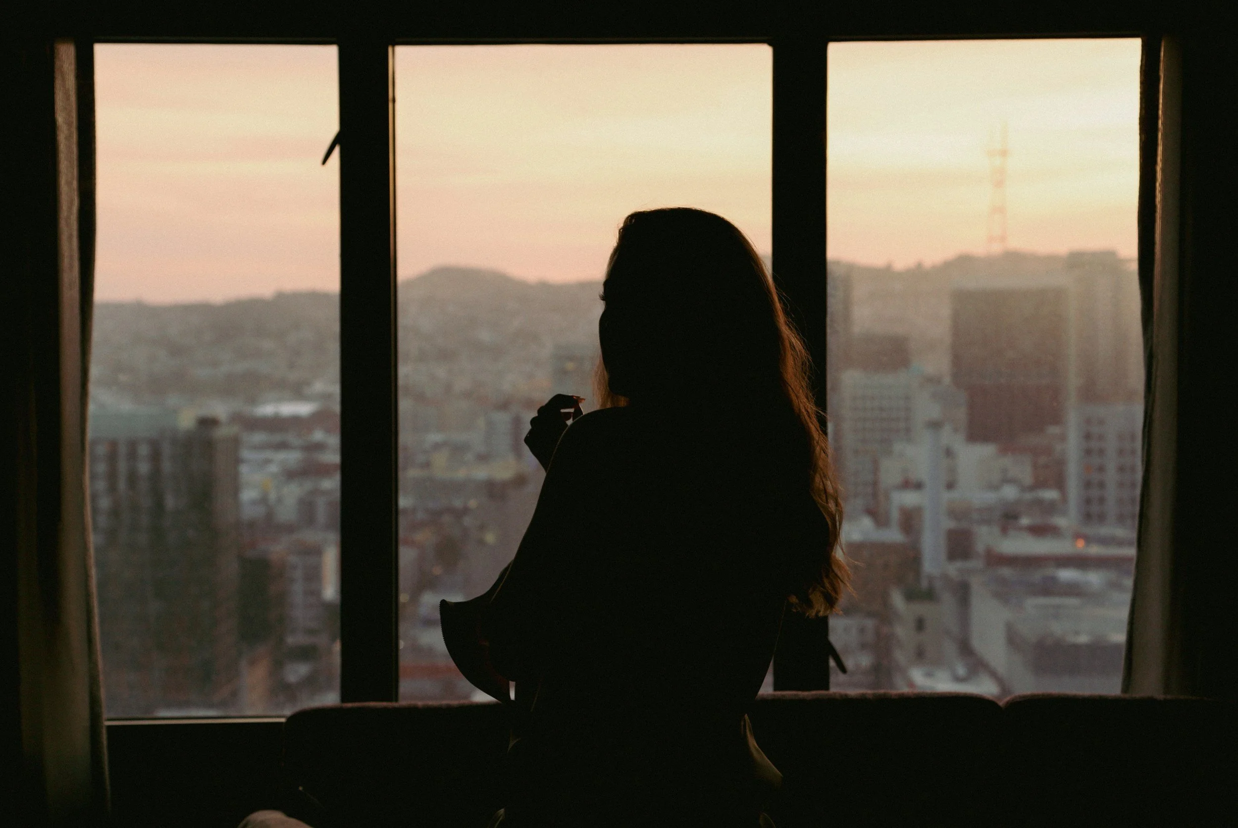 Person gazing out a window at dusk, sitting with the feeling of being behind in life