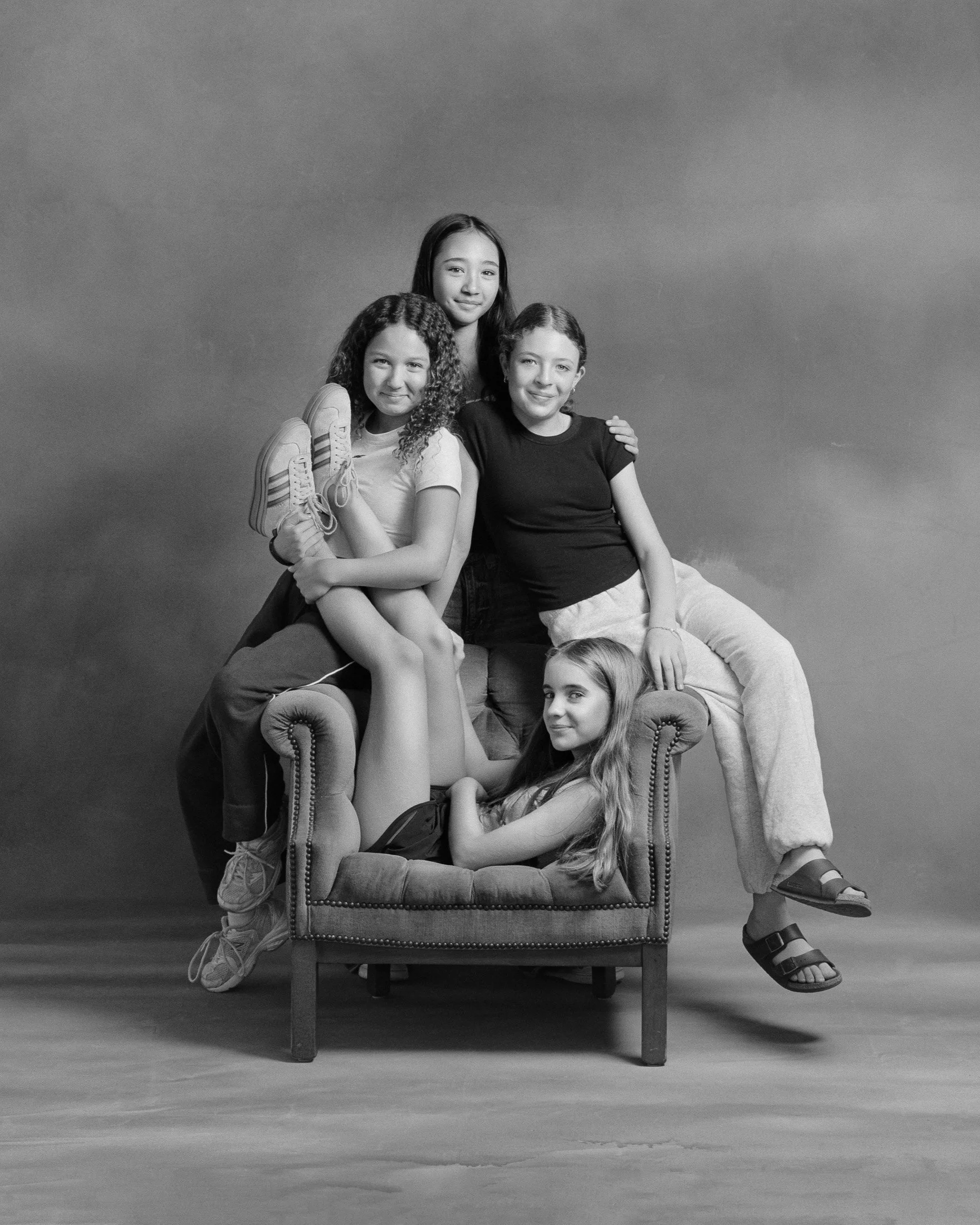 Black and white photo of four young girls posing creatively with a vintage armchair, one seated inside on the cushion, two sitting on it, and one standing behind, all smiling and dressed casually.