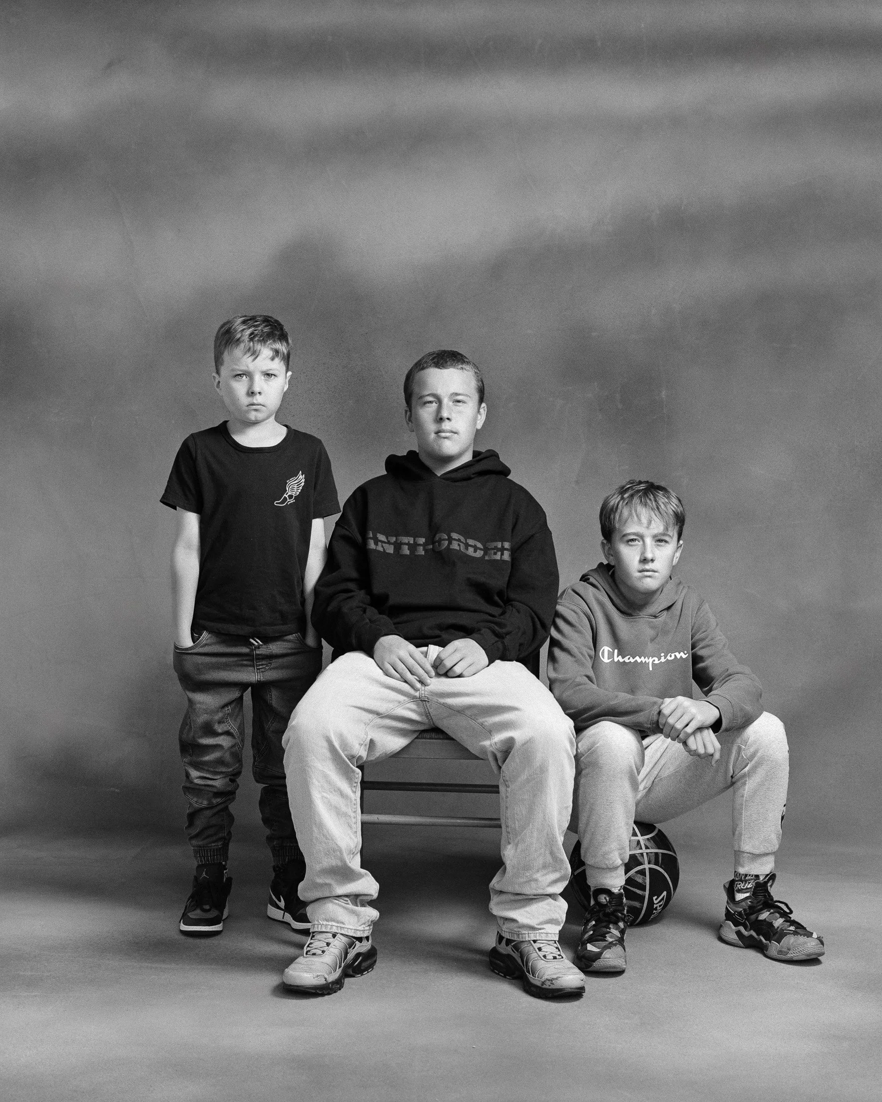 Three young boys in casual clothing posing for a black and white studio portrait. One boy is standing on the left, while the other two are seated; one with hands on knees and the other with hands clasped. The seated boy on the right has a basketball 
