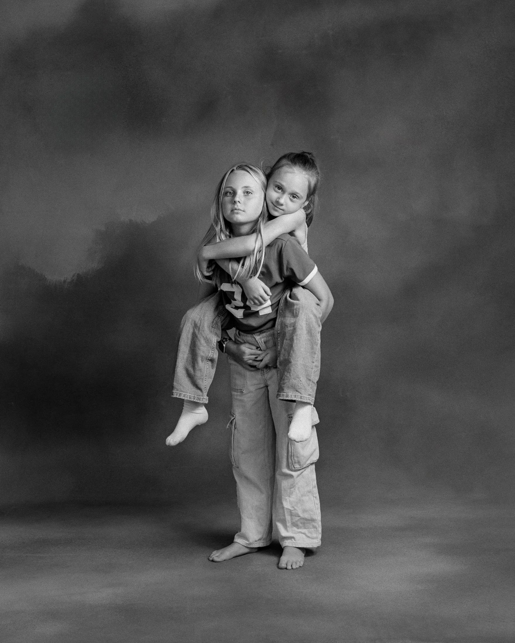 Two young girls, one giving the other a piggyback ride, stand together in a studio with a plain dark background. The girl on the back looks directly at the camera, while the girl carrying her looks slightly side. Both are casually dressed in T-shirts