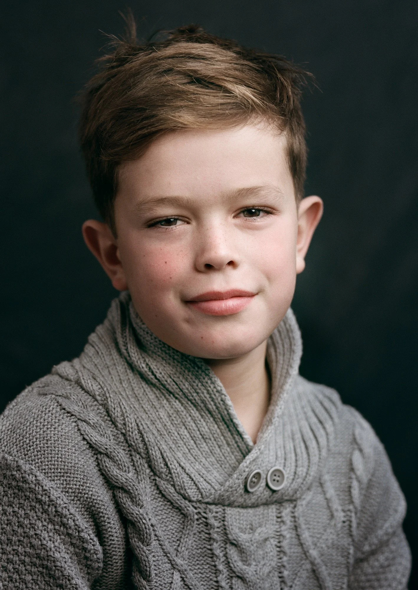 Portrait of a young boy with light skin and short brown hair, wearing a cozy gray cable-knit sweater, against a dark background.