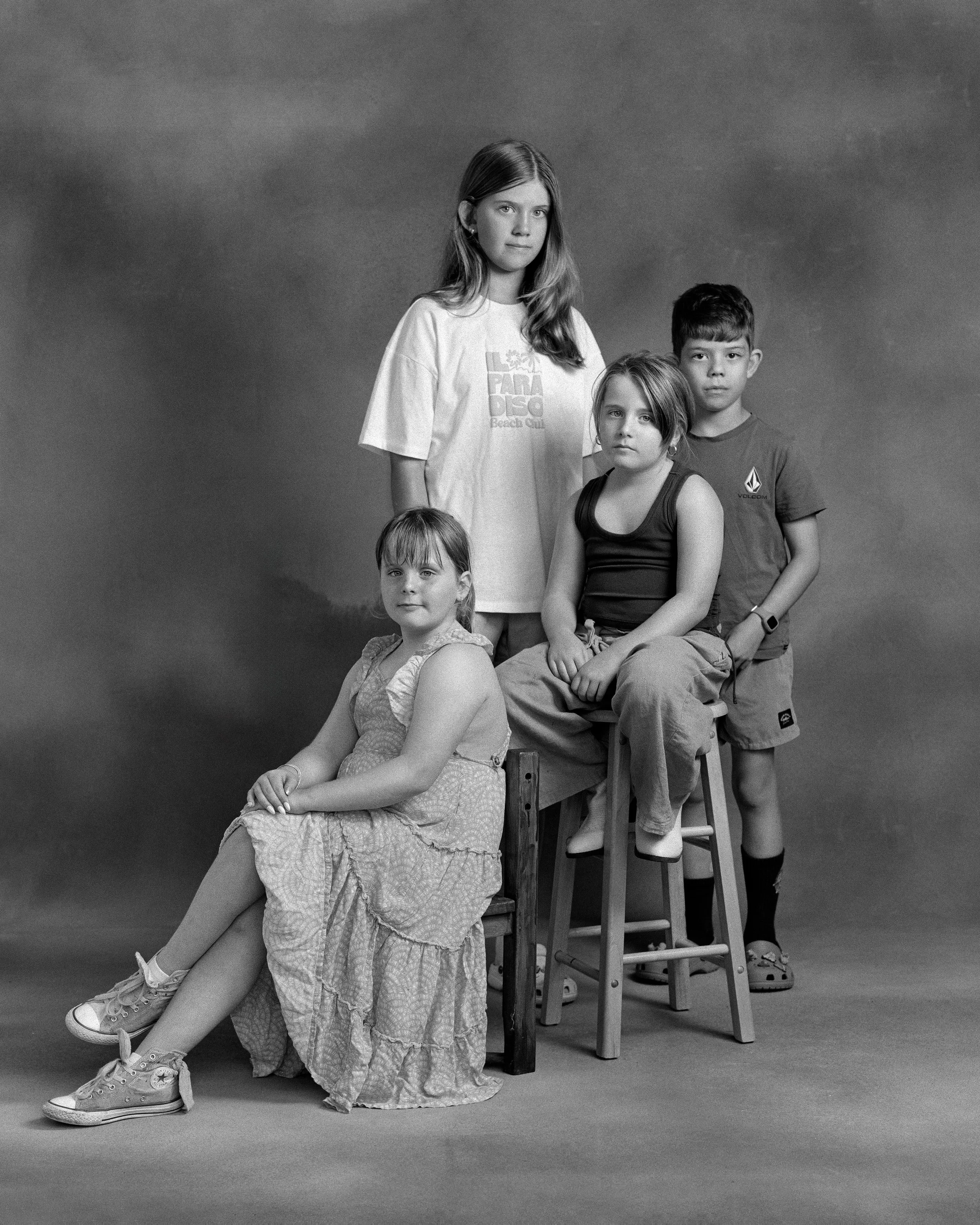 Black and white photo of five children posing for a studio portrait against a plain background. Two girls are seated, and three children are standing behind them.