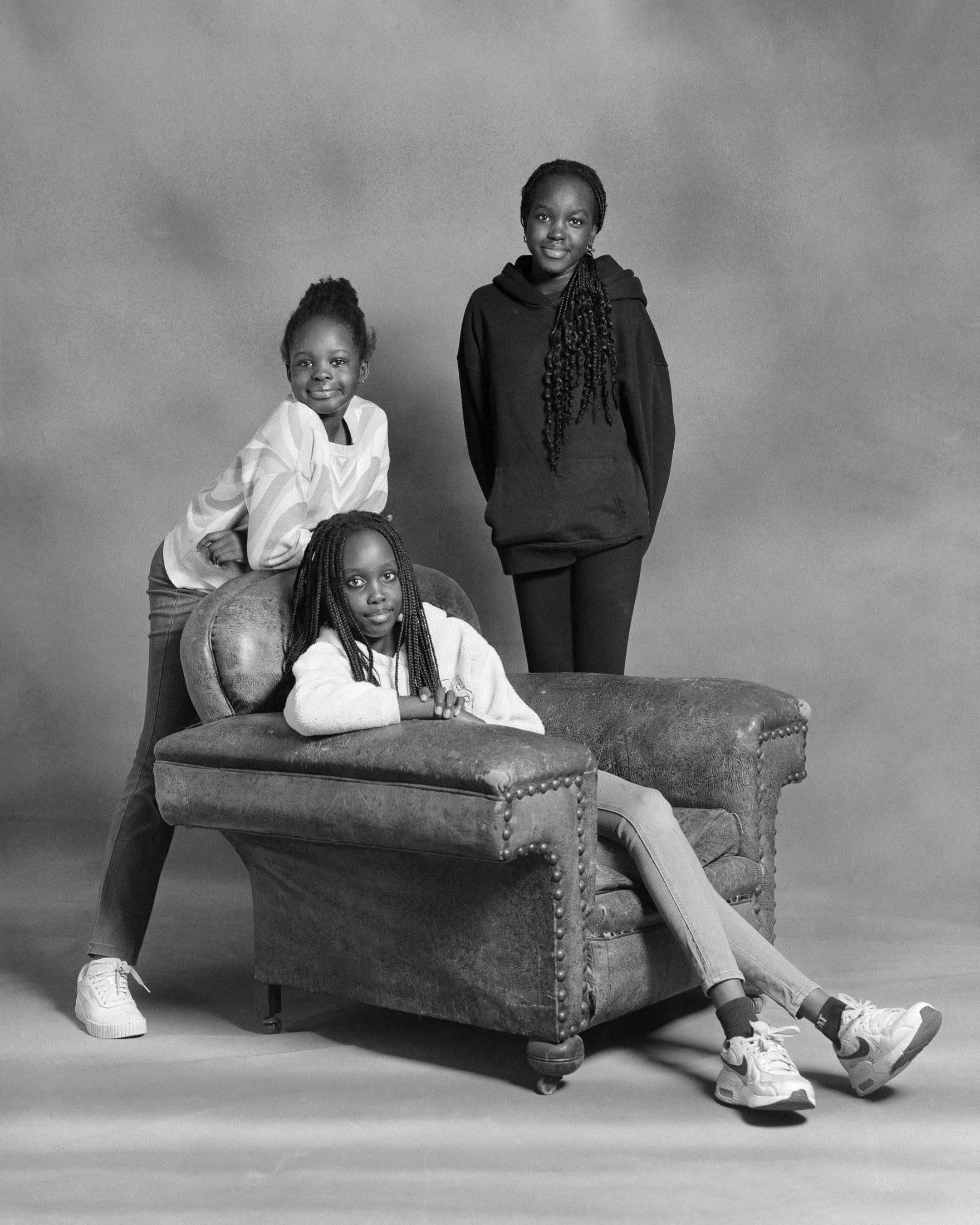 Three young girls with braided hair posing with one sitting on a vintage armchair and two standing behind it in a photography studio, black and white