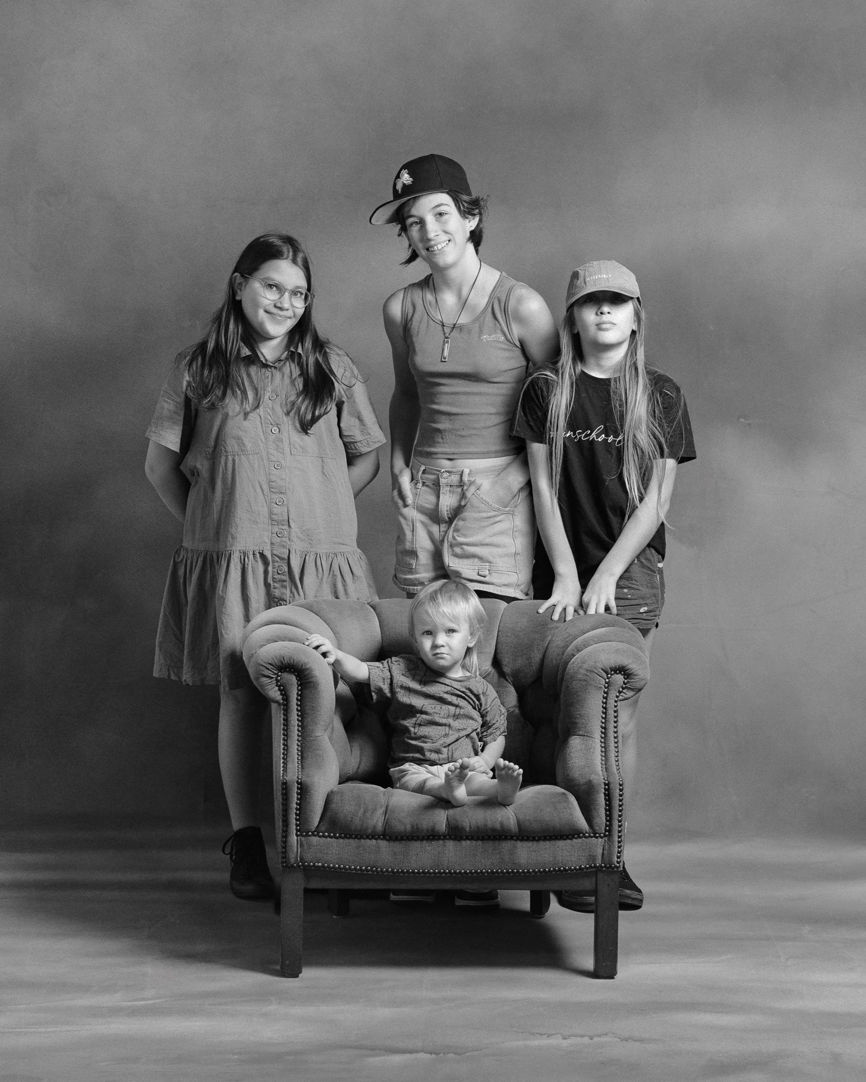 Black and white photo of four girls and a toddler sitting and standing around an armchair against a plain background.