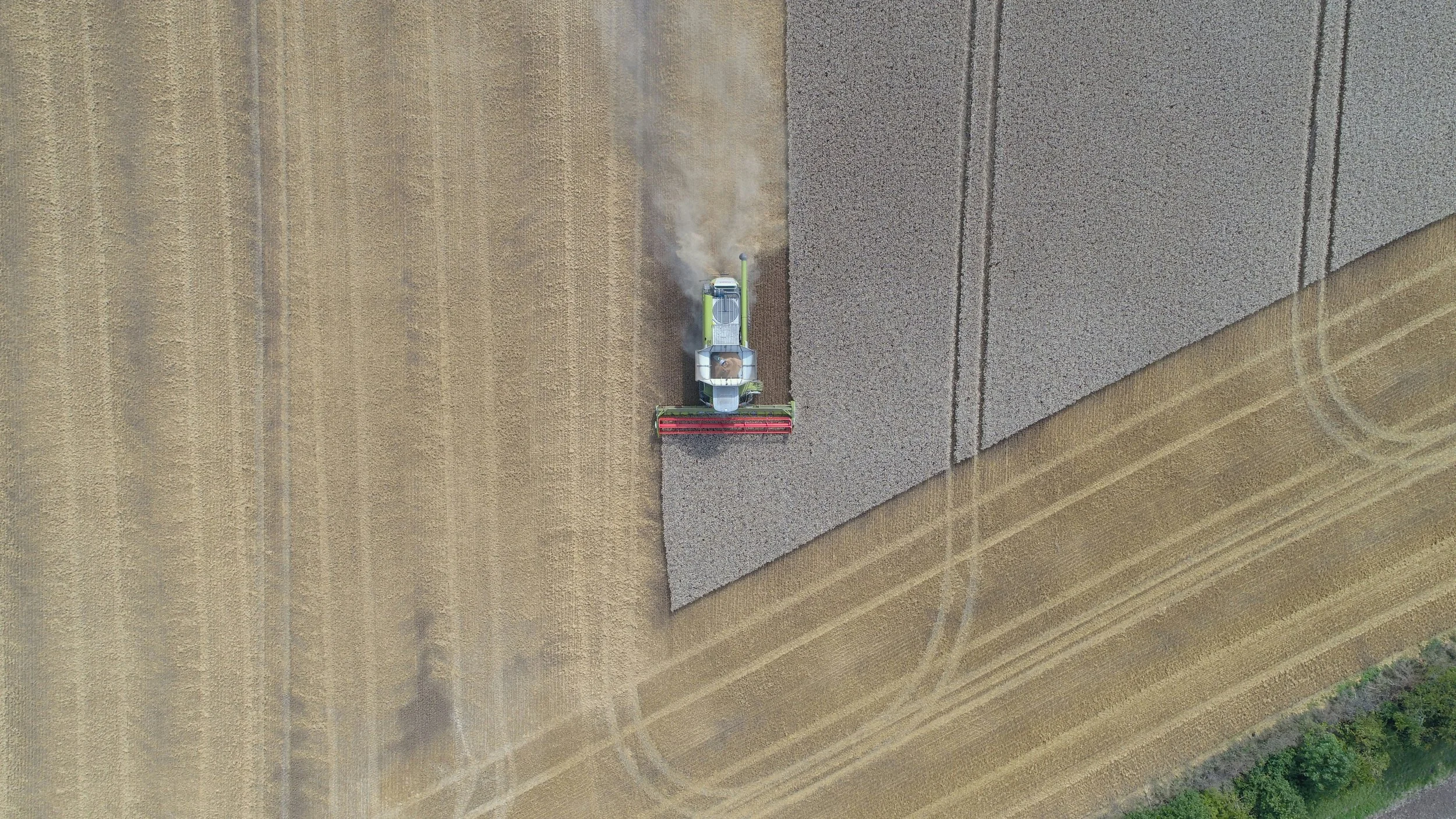 Aerial view of a tractor harvesting a field, with the tractor in the process of collecting crops from the right side and leaving a trail of dust behind.