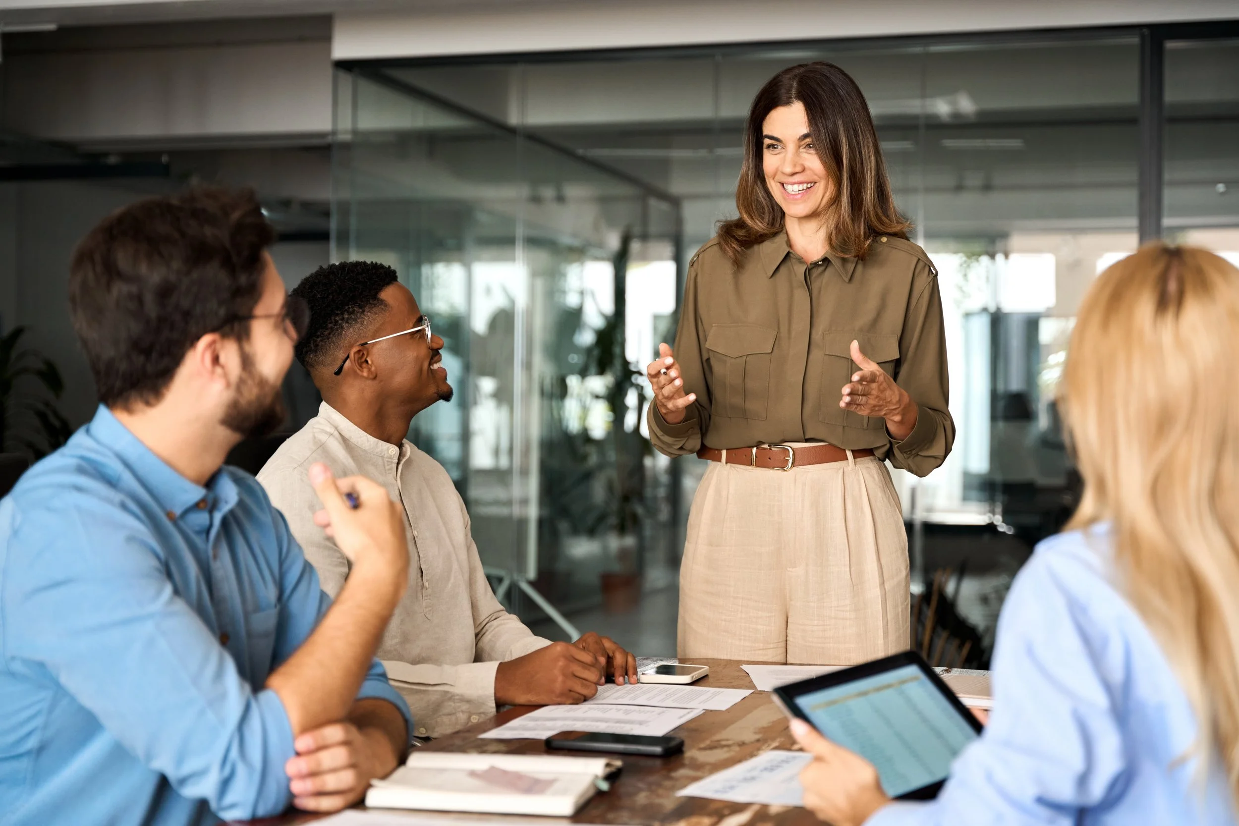 A diverse group of four people are having a meeting in a modern office. A woman is standing and speaking to the others, who are seated around a table. The table has notebooks, papers, and digital devices on it.