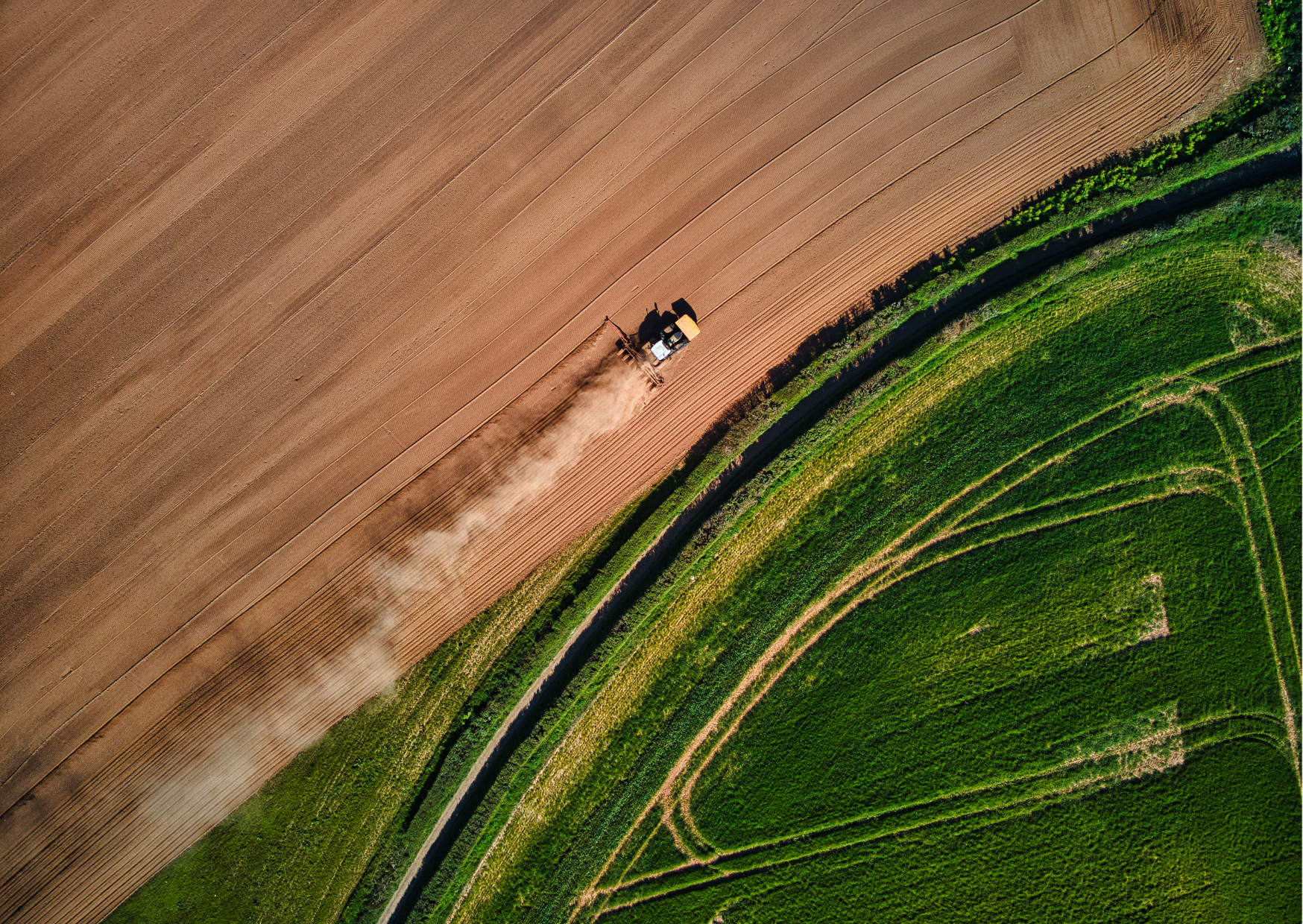 An aerial view of a tractor plowing a field with dust rising behind it, adjacent to patches of green vegetation, separated by a curved black path.