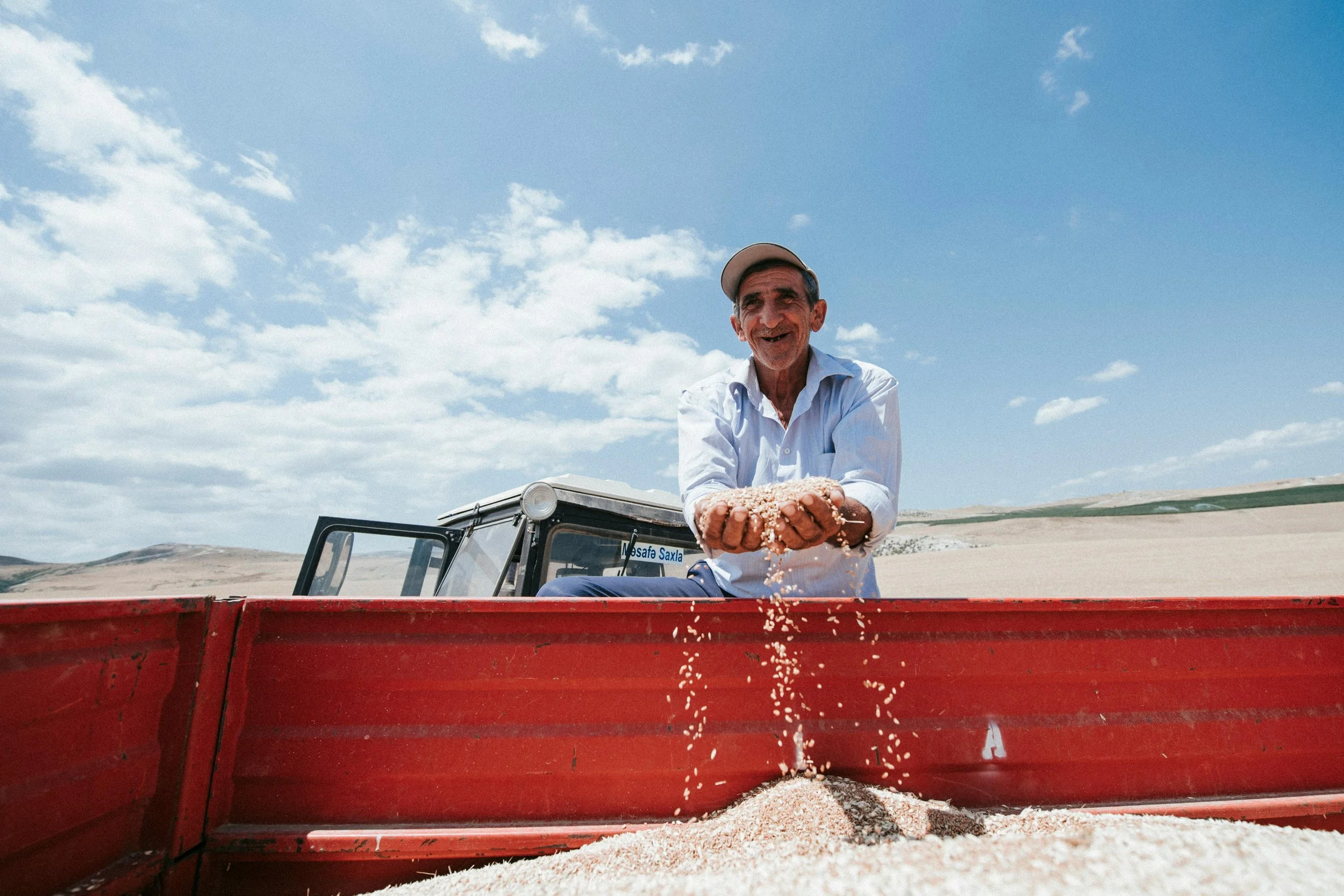 A smiling man in a white shirt and cap pouring grains of rice from his hands into a red container in a rural open landscape under a blue sky with white clouds.