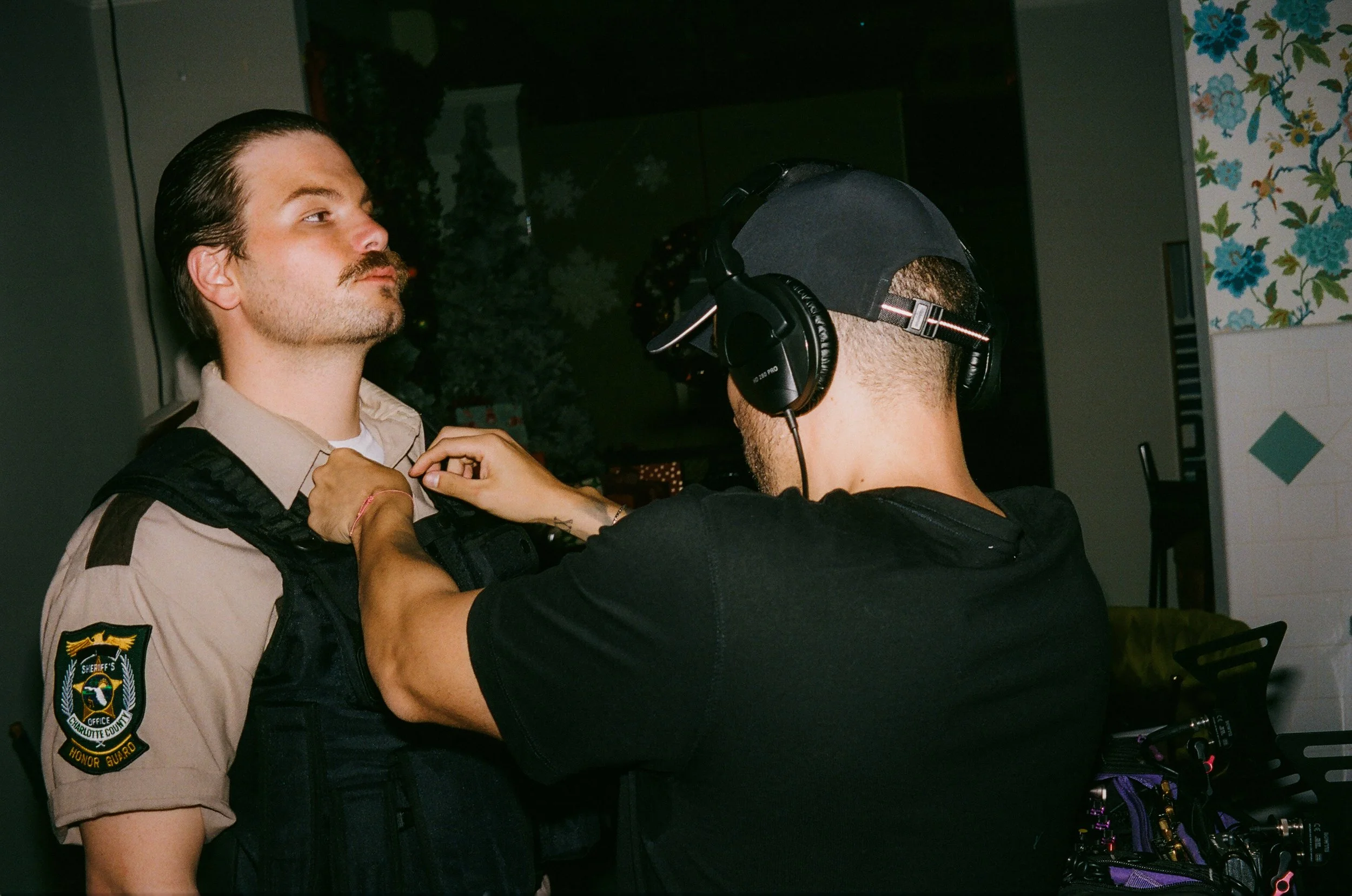 A law enforcement officer wearing a uniform with a Charlotte County sheriff patch is being frisked by a person wearing a black t-shirt and black cap with over-ear headphones. The officer has brown hair, a mustache, and a goatee, and is standing indoo