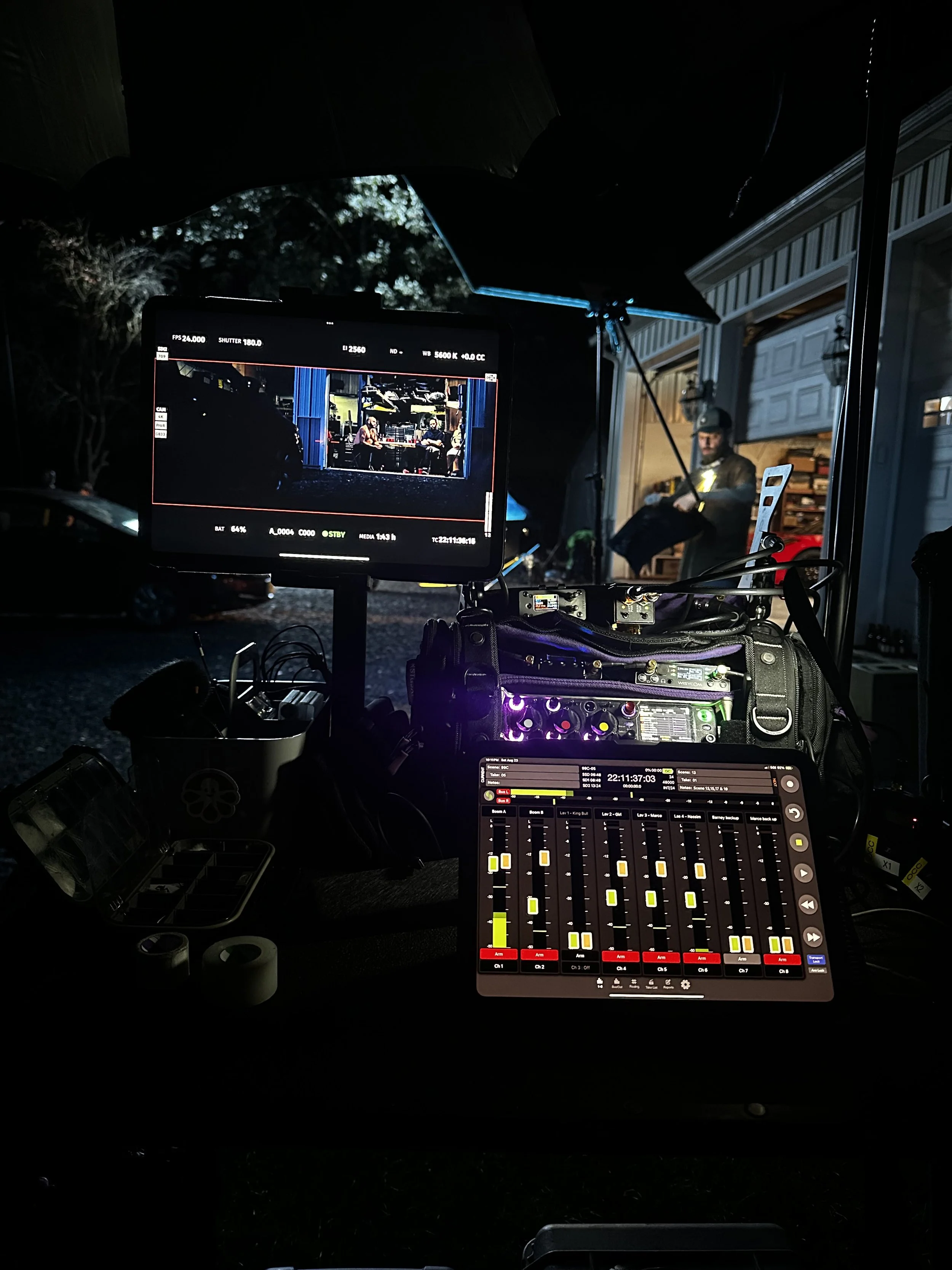Film production set outdoors at night with a monitor showing two people sitting at a table, and a crew member standing nearby under a large umbrella, surrounded by equipment.