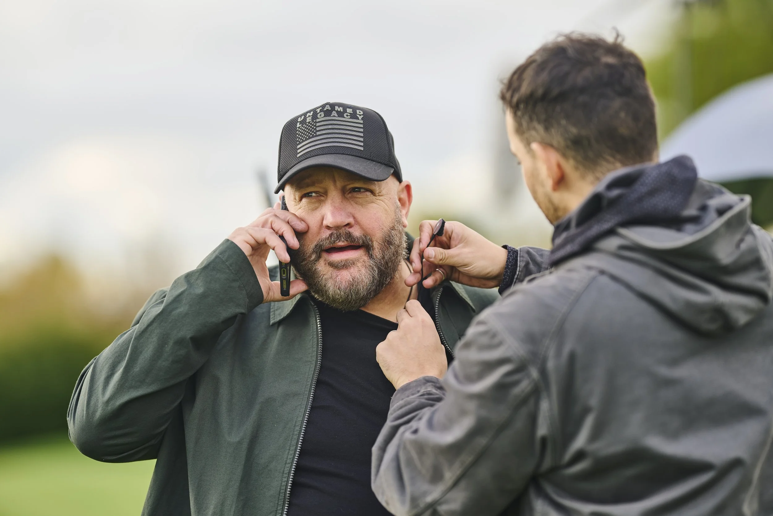 A bearded man wearing a black cap with a U.S. flag and the word 'Military' on it is on a phone call, with a person holding a microphone or recording device close to him. The scene appears to take place outdoors with blurred greenery in the background