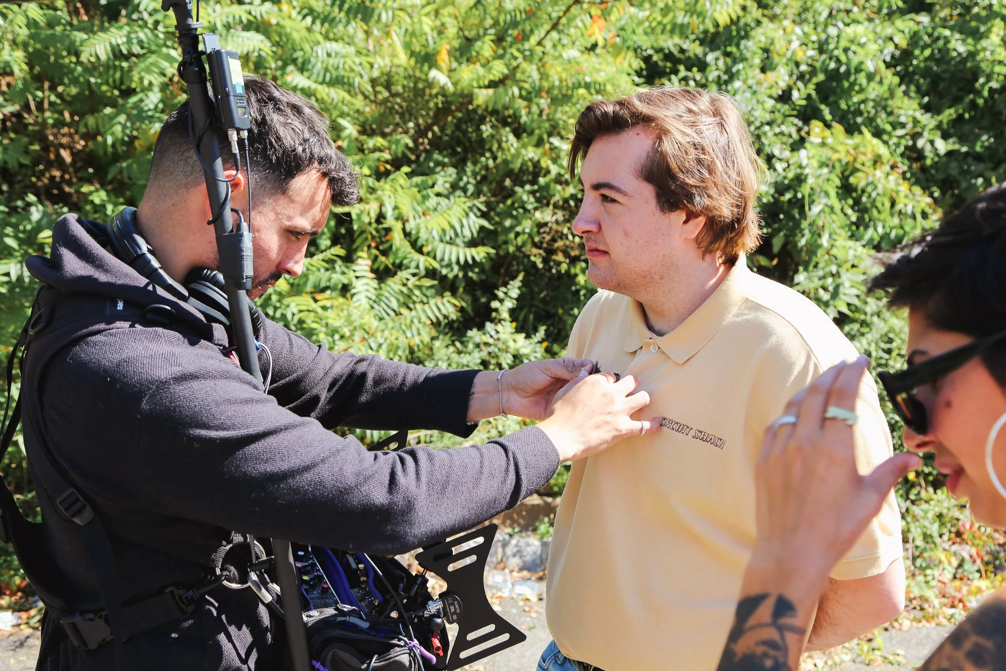 A young man in a yellow polo shirt with the words "Rehabilitation Staff" on it looks at a man in a wheelchair with a headset and a harness. An outdoor setting with green foliage is in the background.