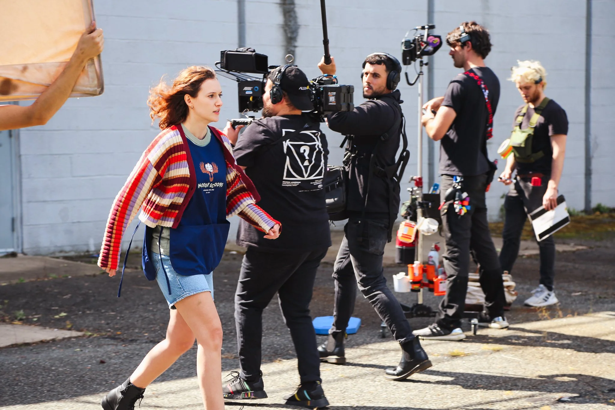 A group of people filming a woman walking outdoors on a sunny day. The woman has brown hair and is wearing a striped jacket over a blue shirt and a denim skirt. The crew is working with cameras, microphones, and other filming equipment.