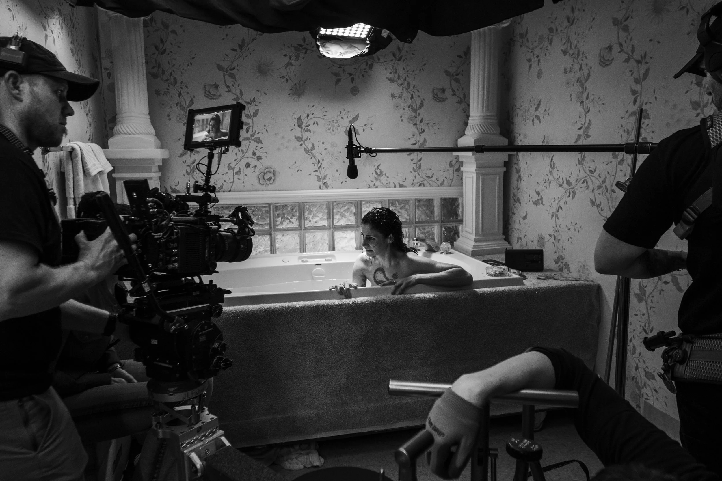 A woman with dark, wet hair is sitting in a bathtub with her arms resting on the edge, surrounded by film crew and cameras recording her in a bathroom with floral wallpaper.