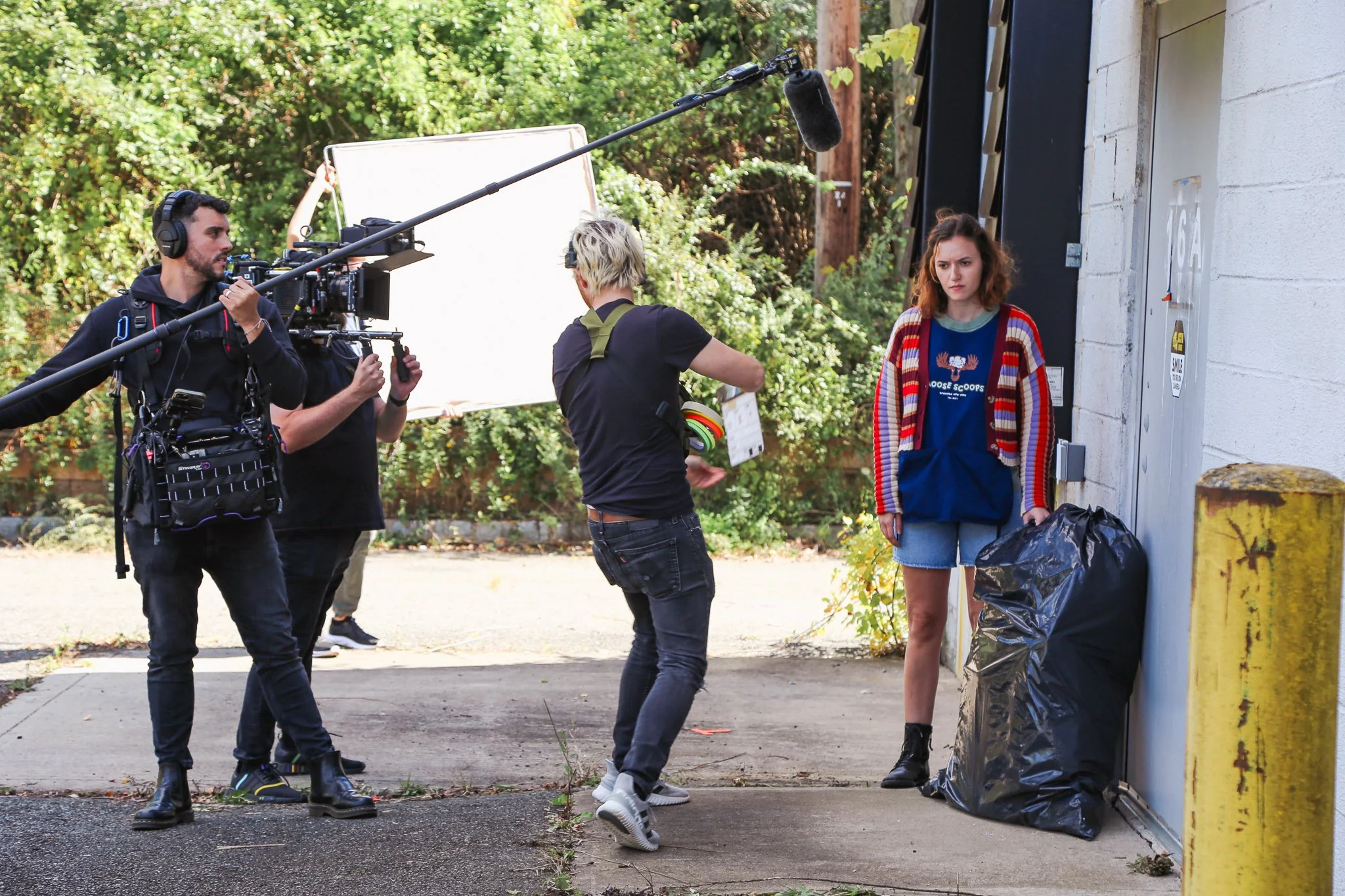 Film crew filming a young woman standing against a wall with a trash bag.