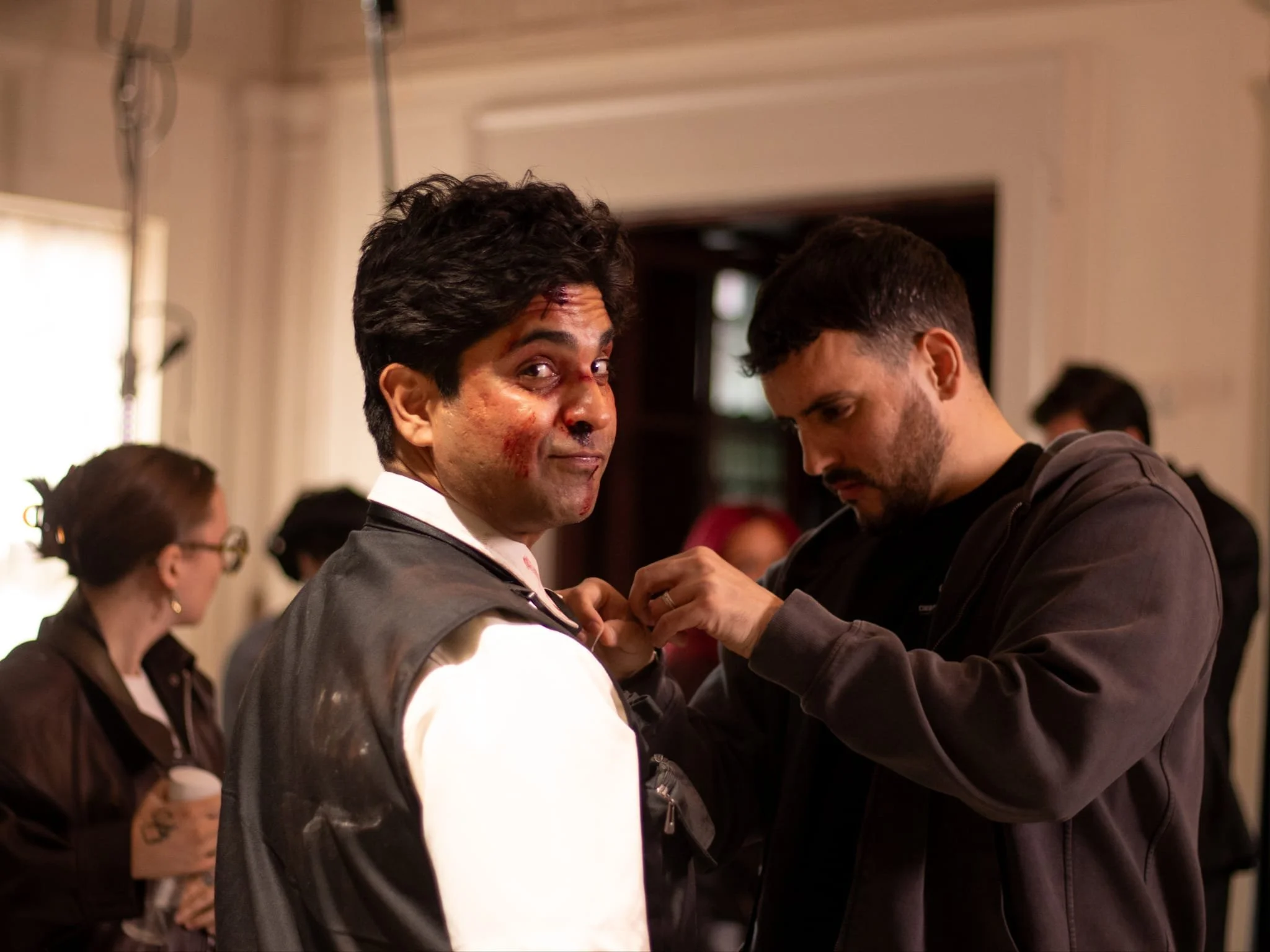 A man with dark, curly hair and a bloodied face turns to look at the camera while another man, with short hair and a beard, assists him with his jacket. In the background, other people are standing in an indoor setting.